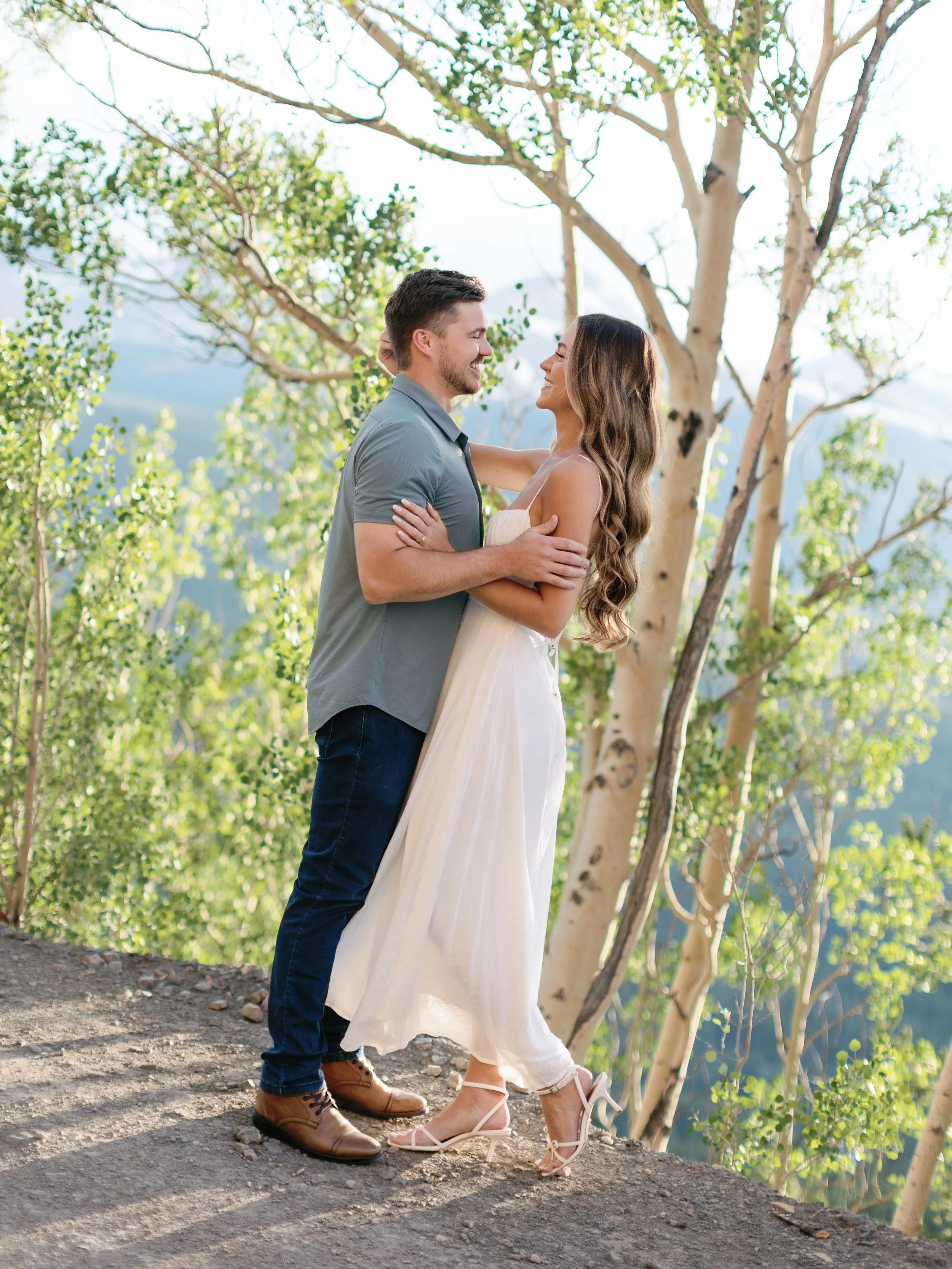 A couple embracing outdoors on a dirt path surrounded by trees and green foliage, smiling at each other.