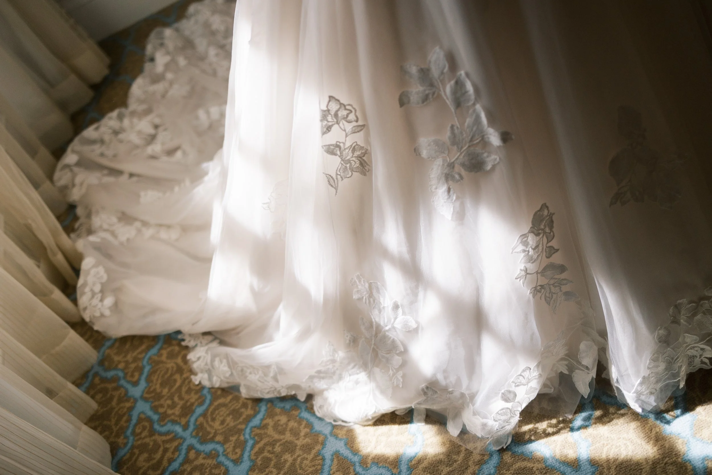 Close-up of a sheer white curtain with floral embroidery, illuminated by sunlight, with a patterned brown and blue carpet underneath.