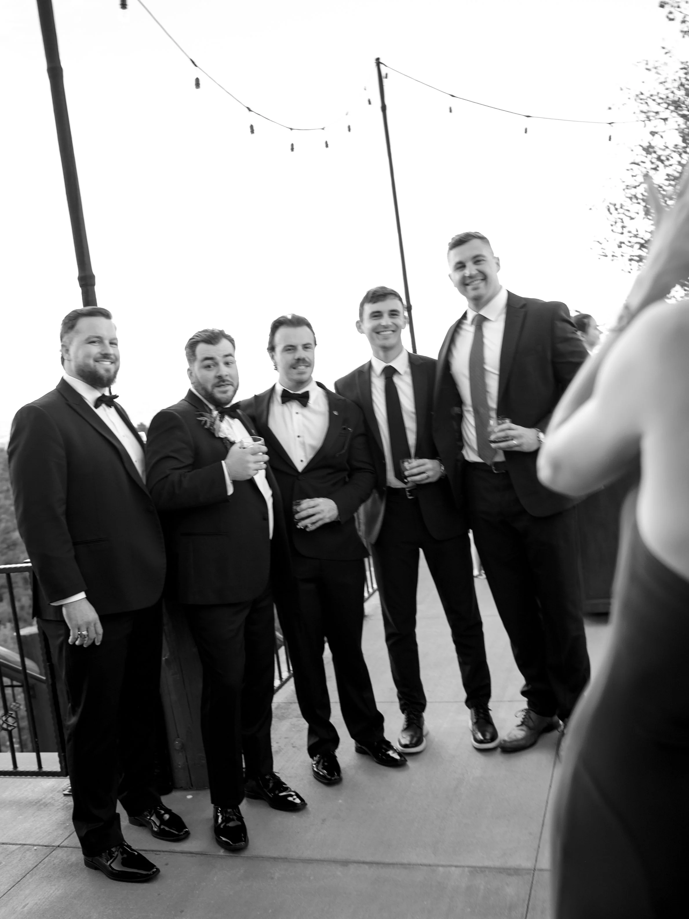 Black and white photo of five men dressed in suits and tuxedos at an outdoor event, standing in a line and smiling at the camera, with string lights overhead.