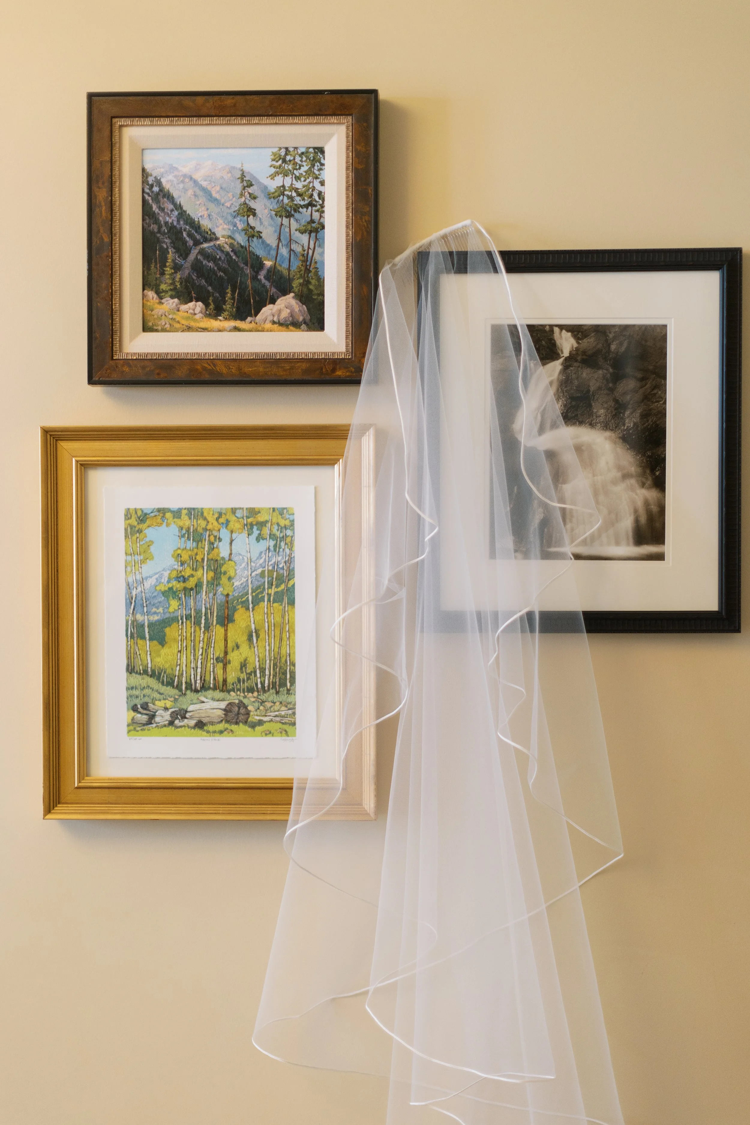A wedding veil hanging on a wall art arrangement with three framed landscape pictures, including mountains, trees, and a waterfall.