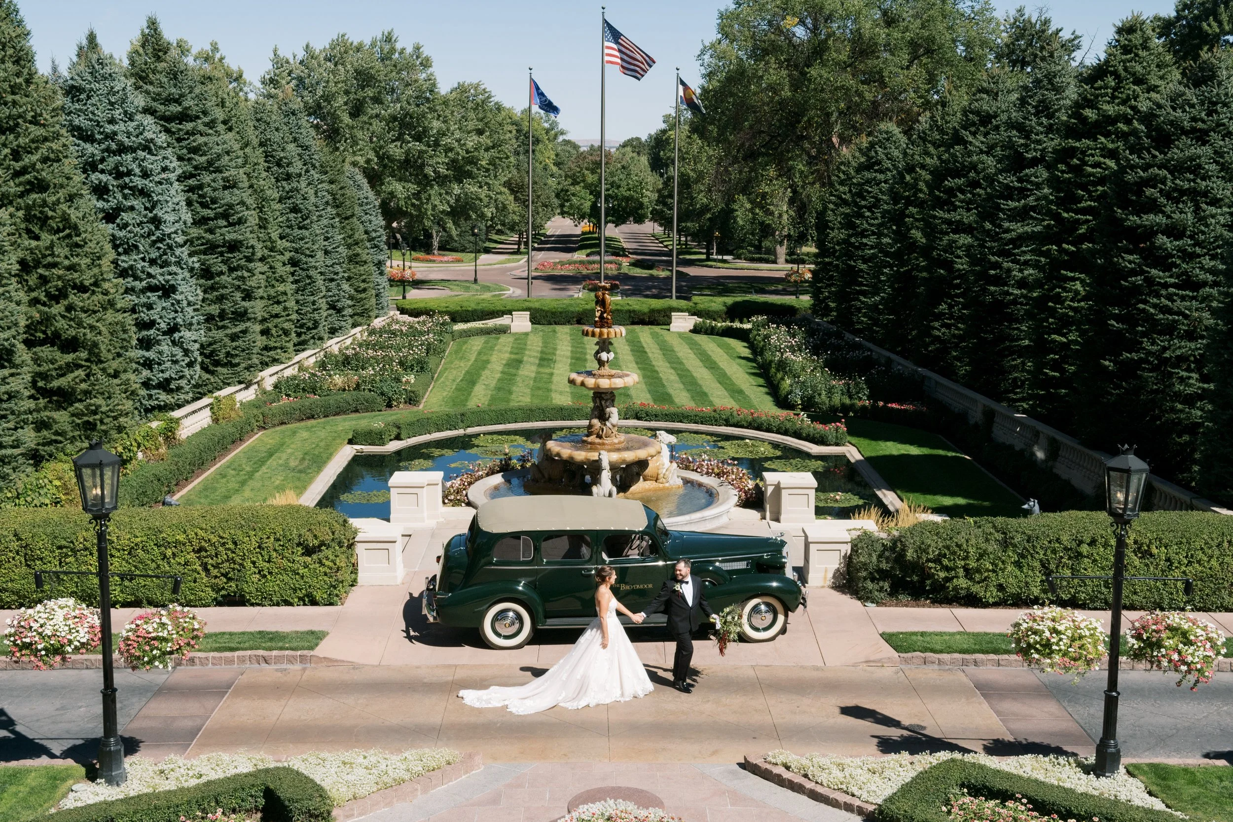 Bride and groom holding hands in front of a vintage car near a fountain with a garden background.