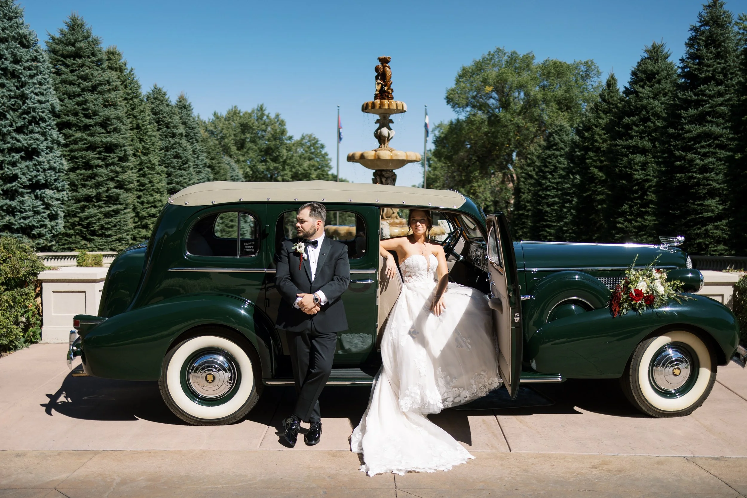 Bride in white wedding dress sitting in a vintage green car, groom in black tuxedo standing outside the car, in front of a fountain and trees.