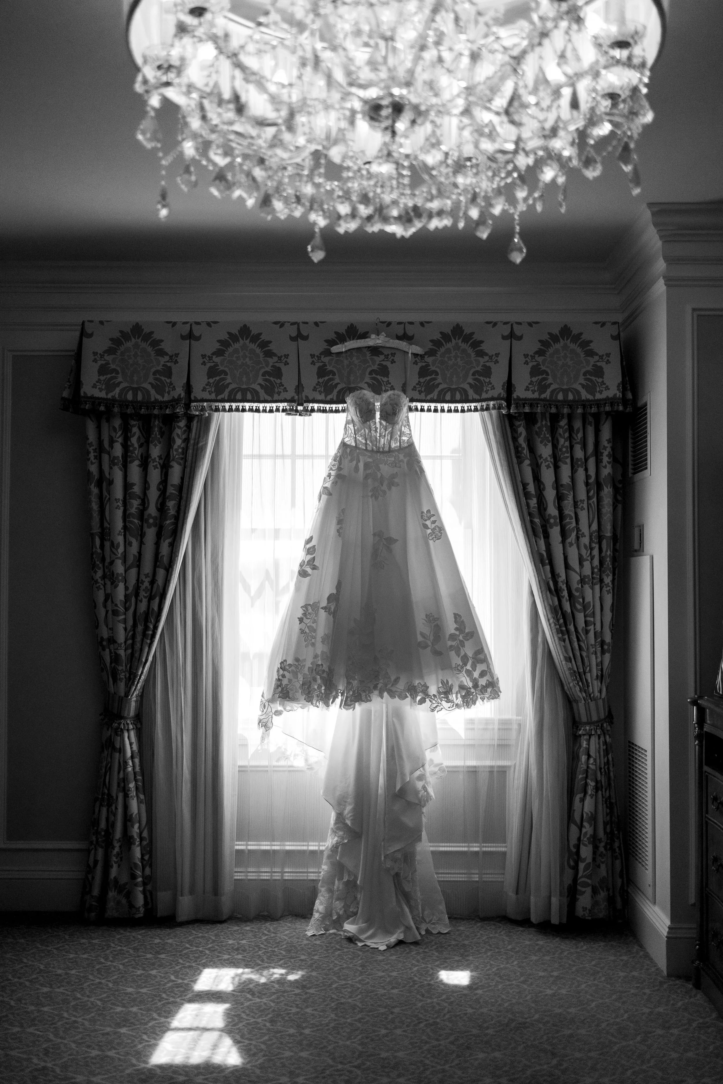 A wedding dress hanging in front of a window with curtains, illuminated by sunlight, with a chandelier above and patterned carpet below.