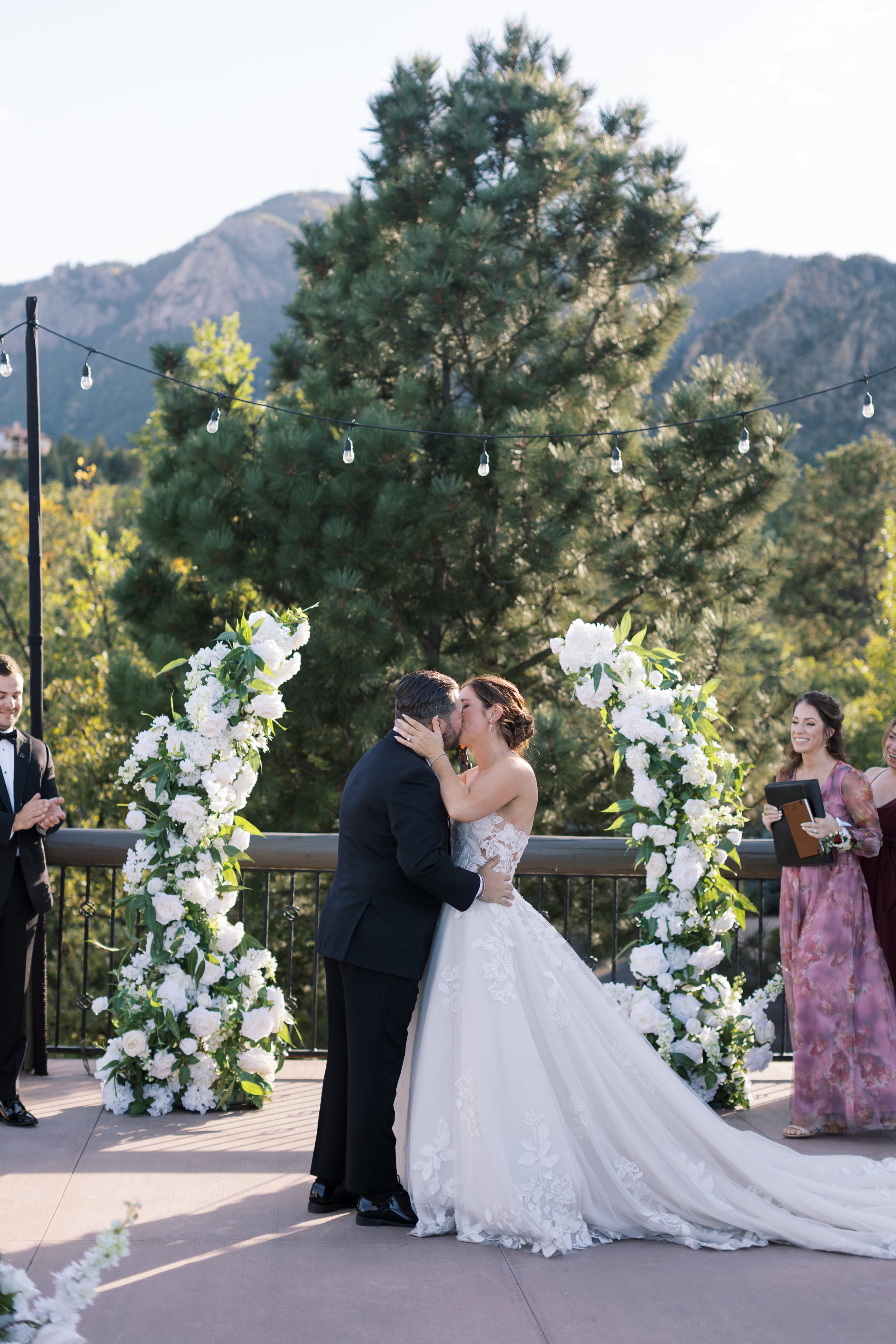 A couple in wedding attire kissing during their outdoor wedding ceremony underneath a large tree, with mountains in the background and floral decorations surrounding them.