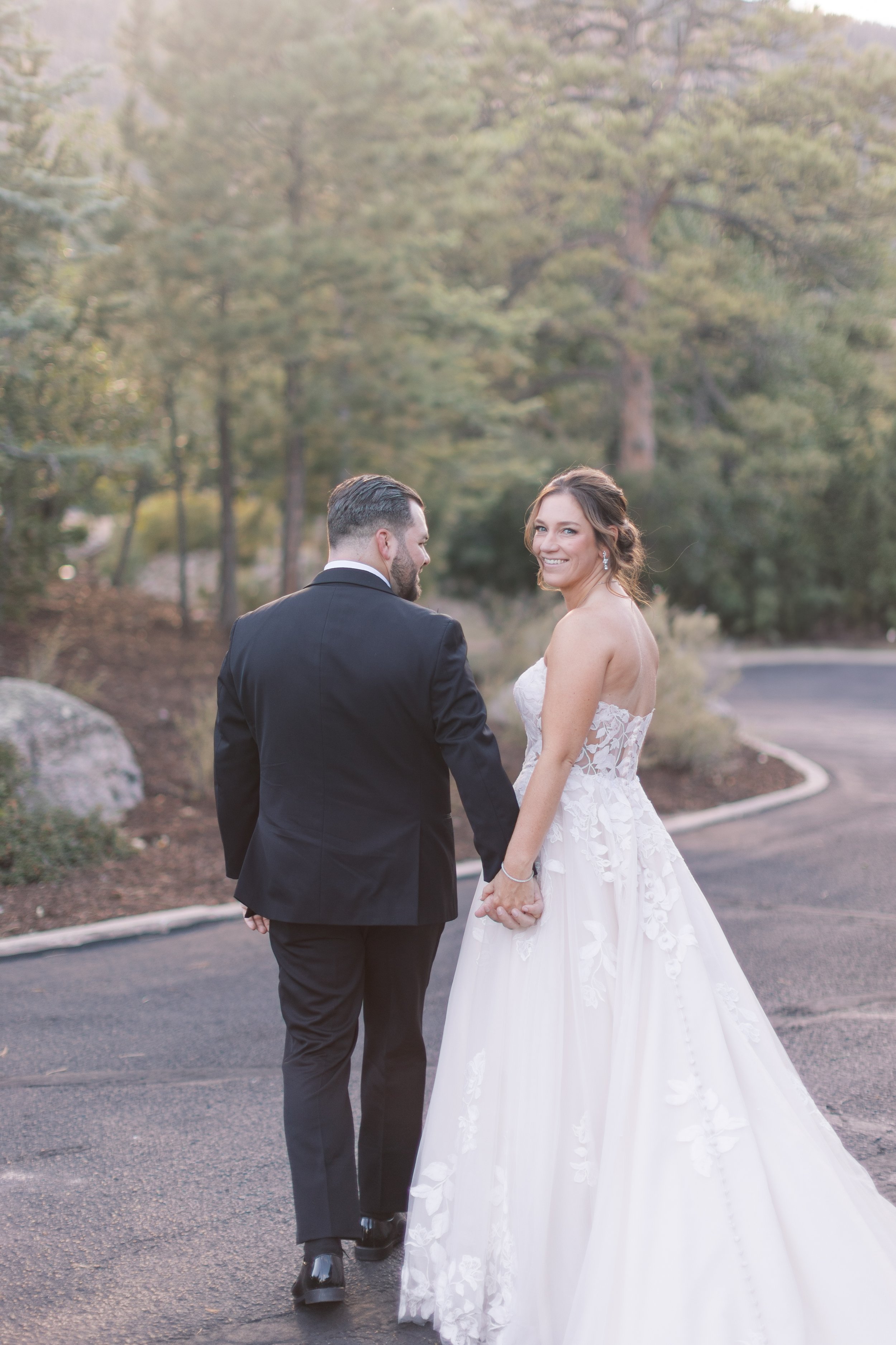 A bride and groom walking hand-in-hand outdoors during a wedding, with trees and a curved road in the background.