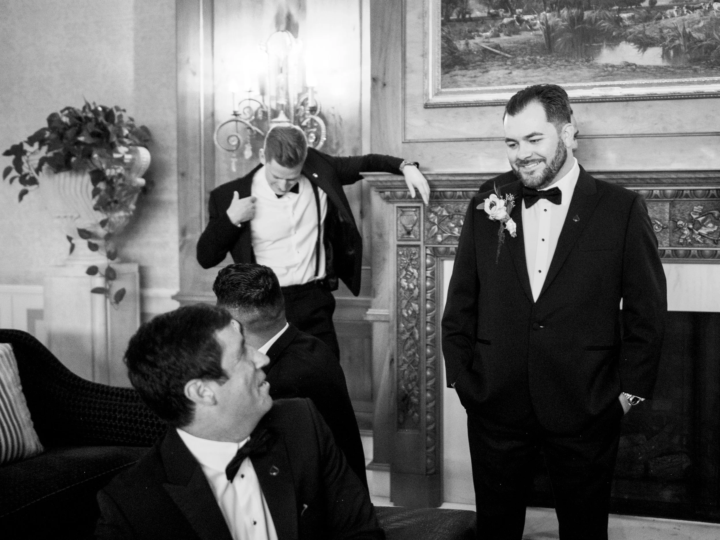 Four men dressed in tuxedos, two sitting and two standing, in an elegant room with a fireplace, ornate wood trim, and a landscape painting, engaging in conversation and smiling.