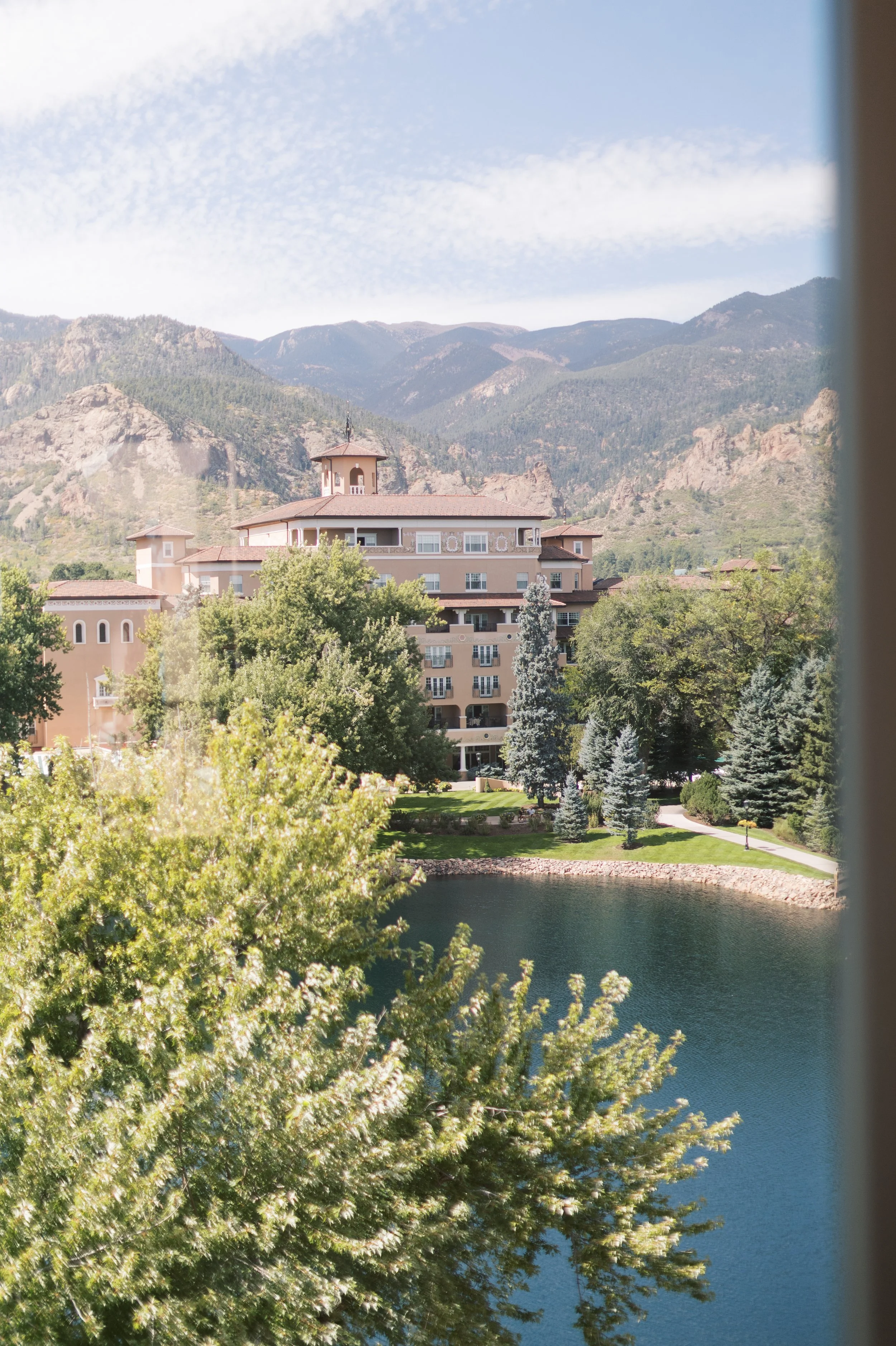 View of a large beige building with multiple stories and balconies, surrounded by green trees, beside a small body of water with mountains in the background under a partly cloudy sky.