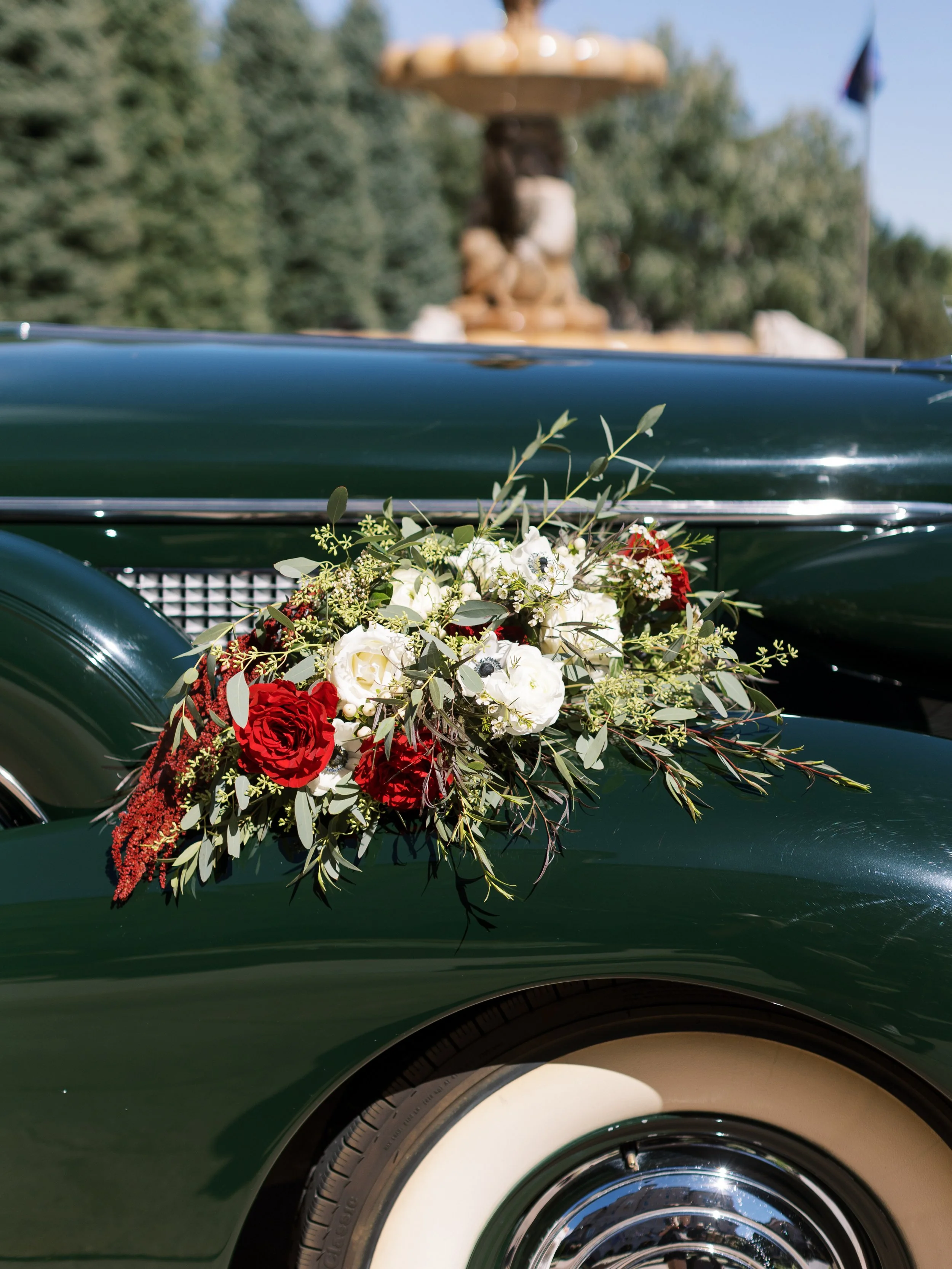 A vintage green car decorated with a floral arrangement of red and white roses, eucalyptus leaves, and other greenery, for a wedding or special event.
