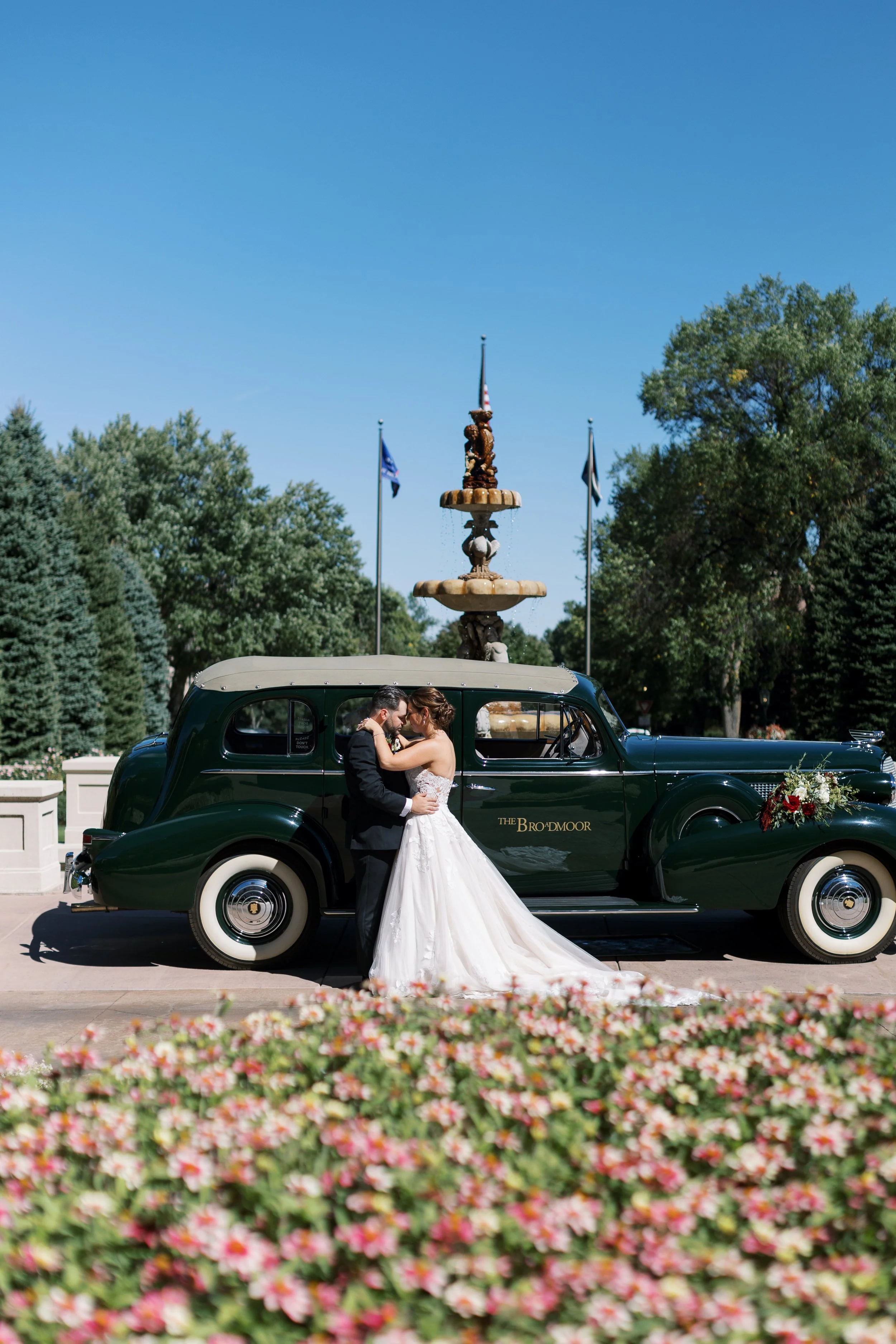 A bride and groom sharing a moment next to a vintage car with a fountain in the background, surrounded by greenery and flowers.
