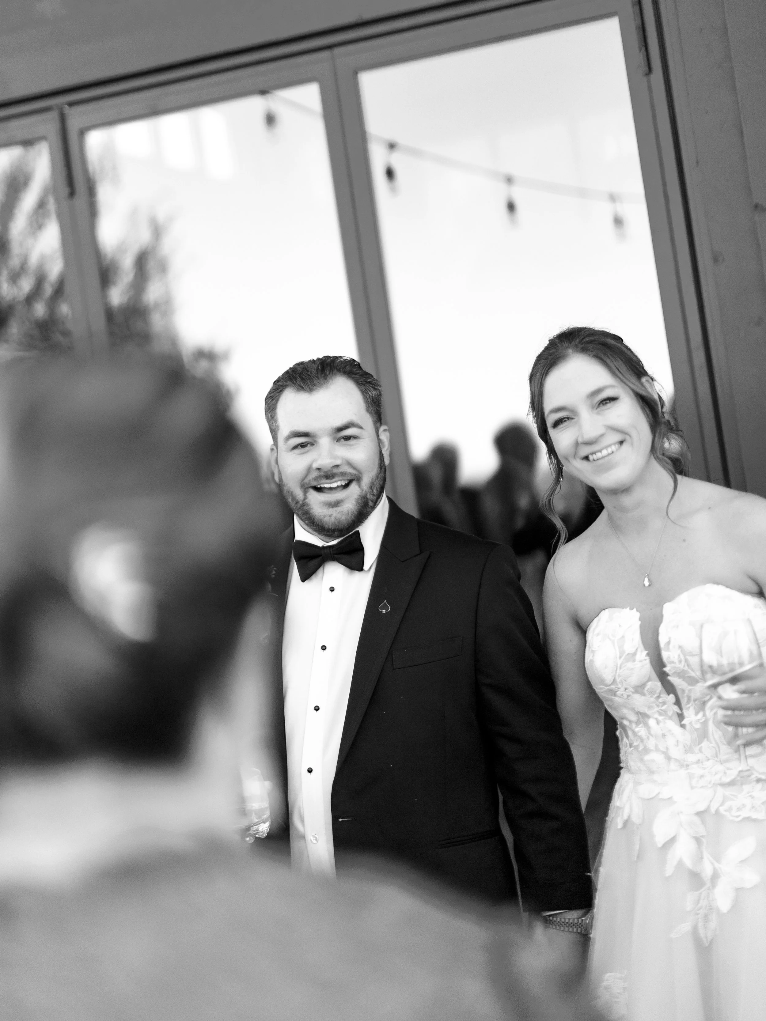 Black and white photo of a smiling bride and groom at their wedding reception, standing near a large window with string lights outdoors in the background.