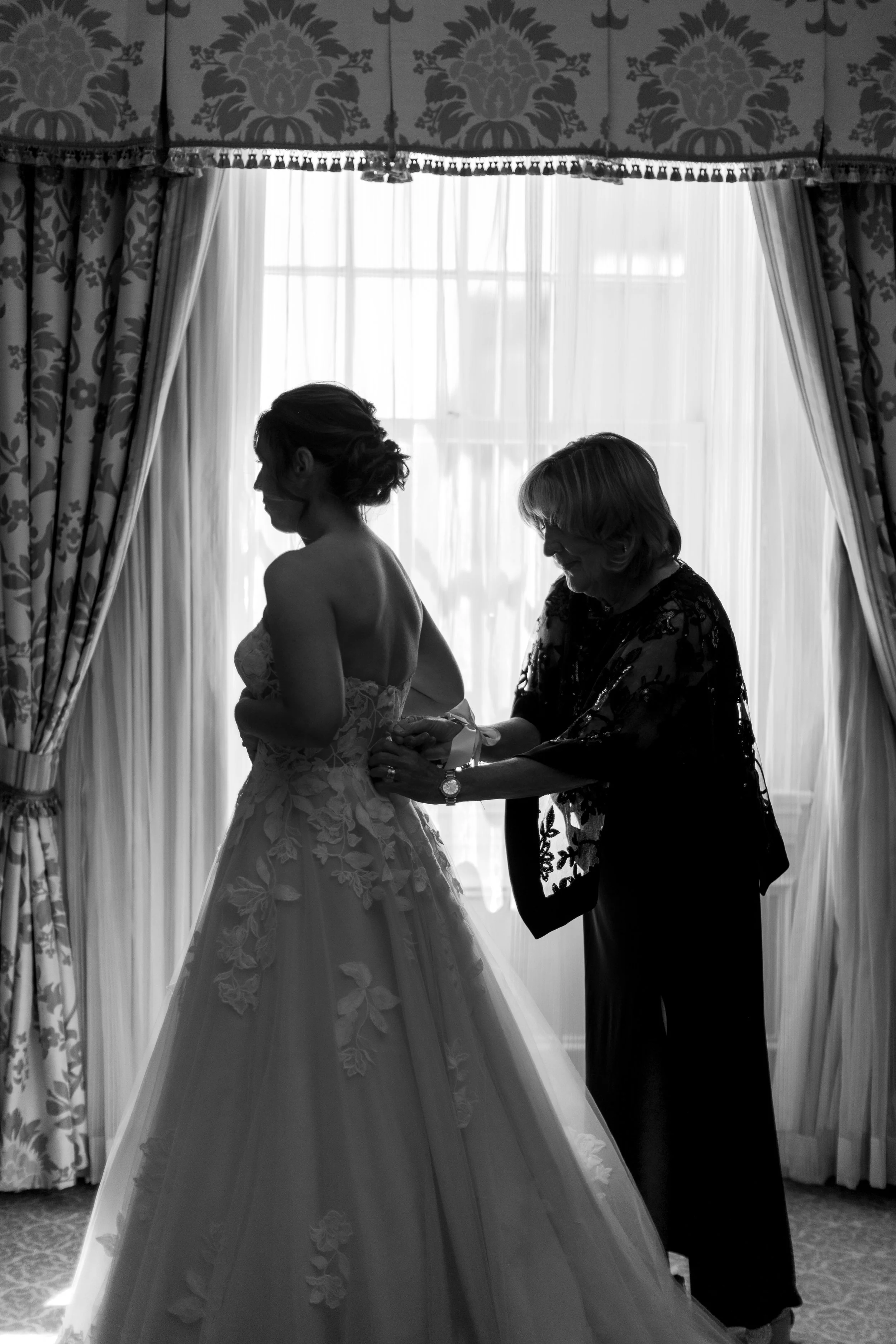A bride in a wedding gown being assisted with her dress by an older woman, possibly her mother, in front of a window with curtains.