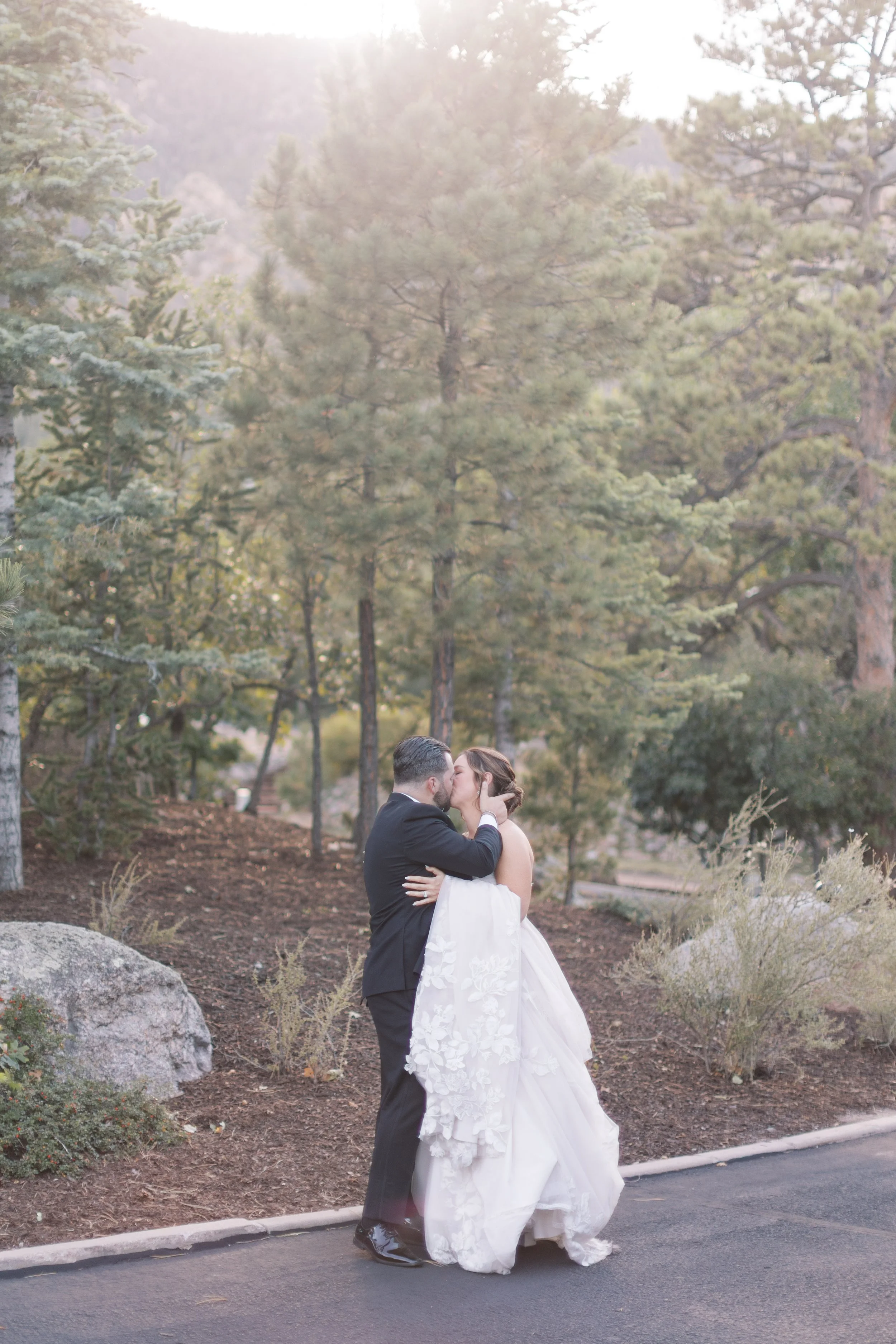A bride and groom sharing a kiss outdoors on a paved path with trees in the background.