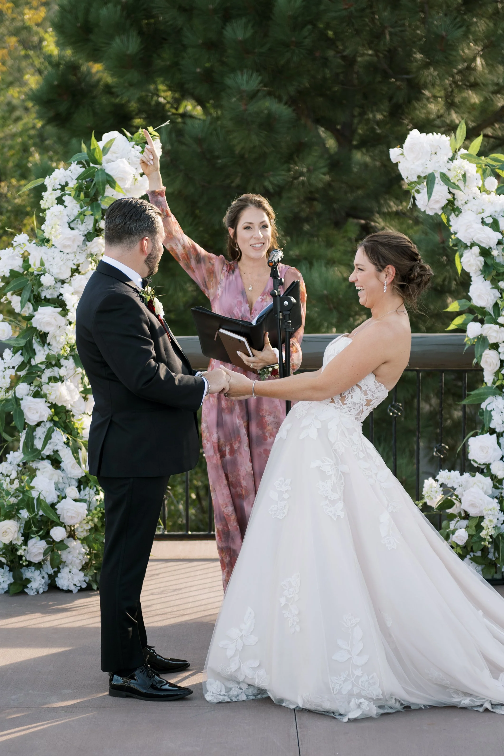A wedding ceremony with a bride and groom holding hands and exchanging vows, officiated by a woman standing under a floral arch outdoors surrounded by greenery.