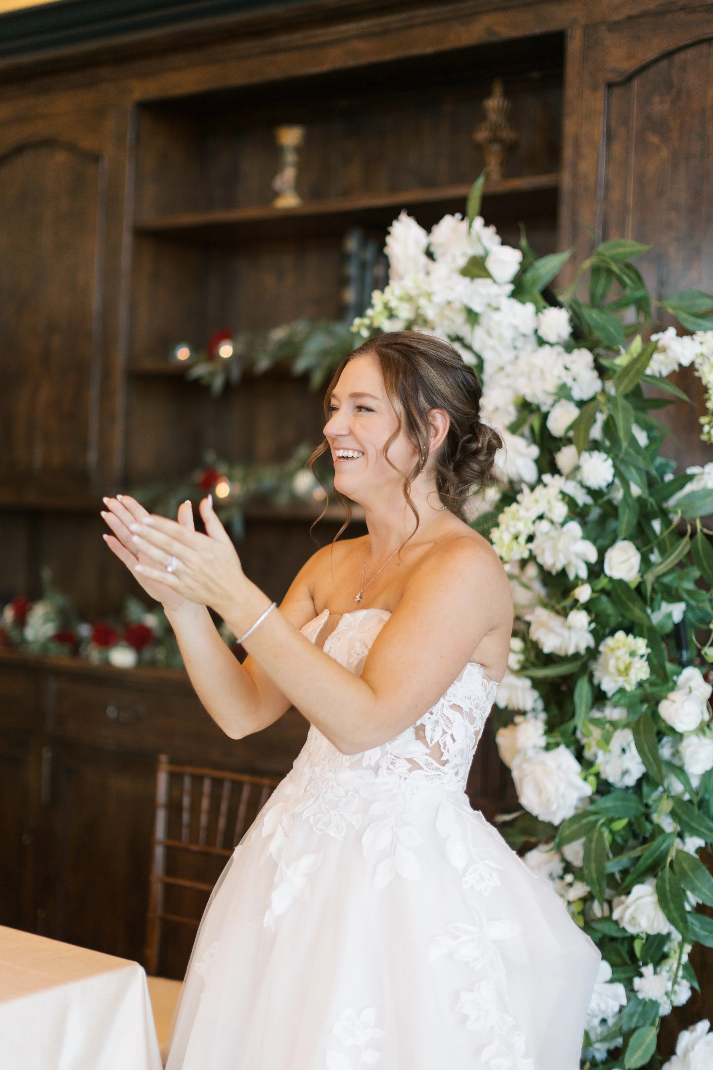 A bride in a strapless wedding gown with lace detail, smiling and clapping, standing near a floral arrangement of white flowers and greenery indoors.