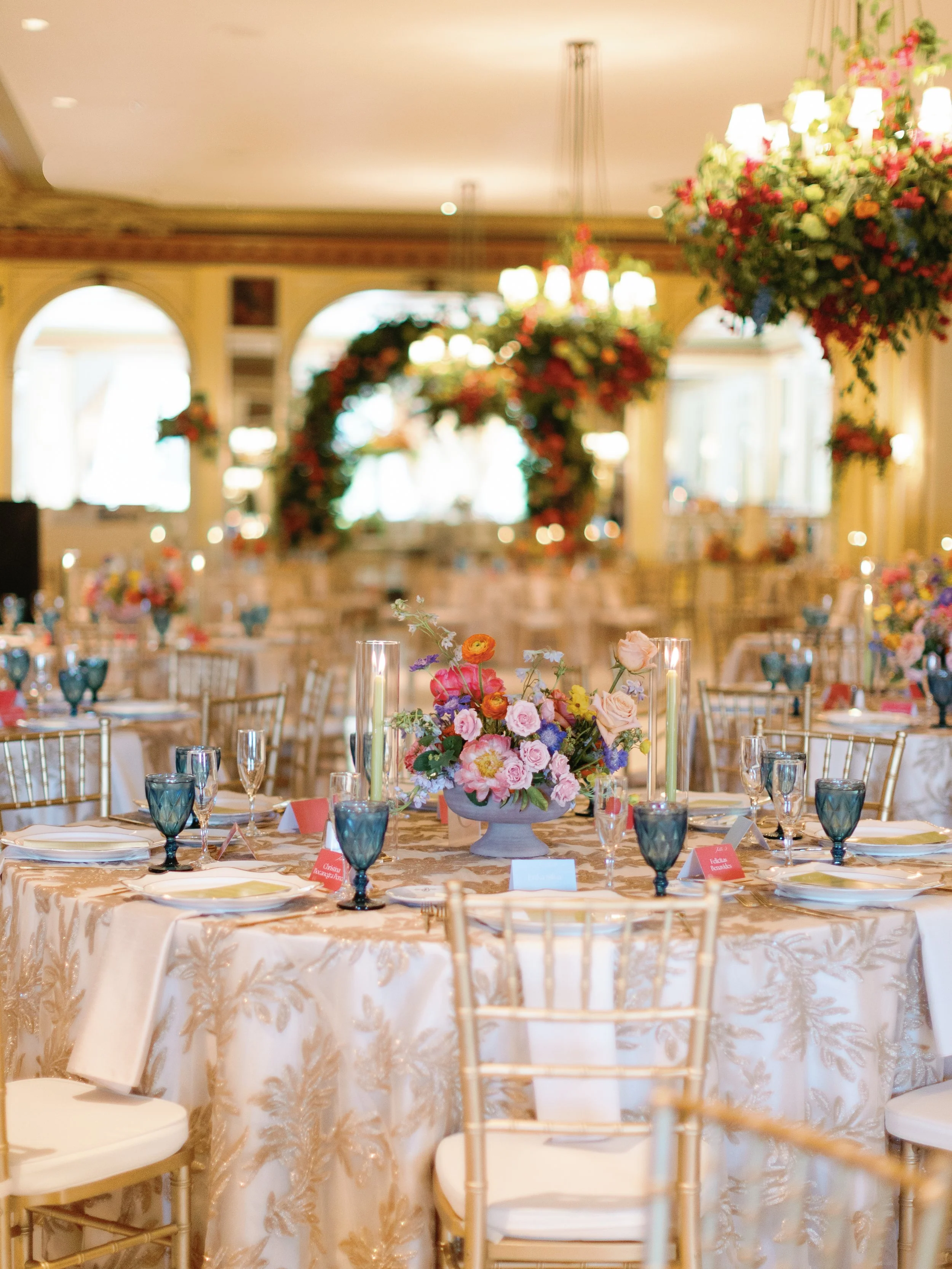 Elegantly decorated banquet hall with round tables covered in embroidered tablecloths, floral centerpieces, and candleholders, with a floral arch and chandelier lighting in the background.
