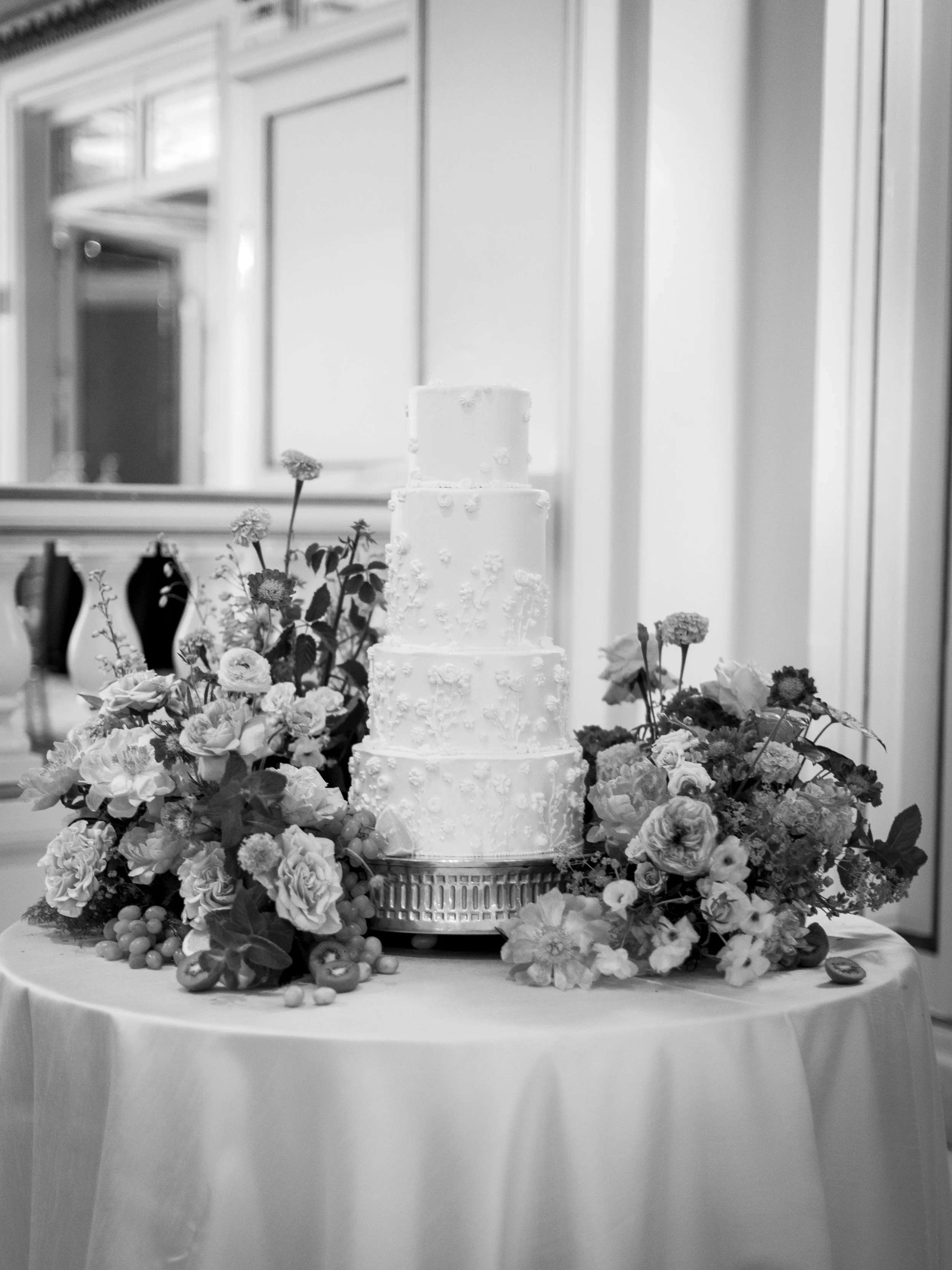 Black and white photo of a four-tiered wedding cake with intricate icing designs on a round table, decorated with an arrangement of flowers and fruit, in a room with paneled walls and a mirror.