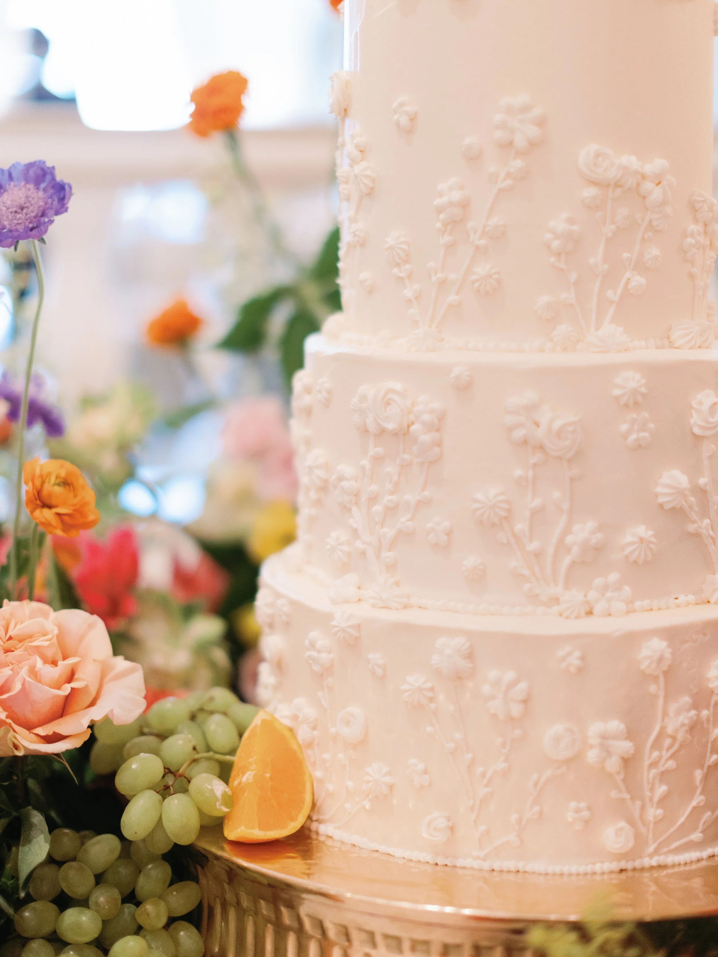 A white, tiered wedding cake decorated with small, delicate floral patterns. The cake is surrounded by colorful flowers, green grapes, and a slice of orange.