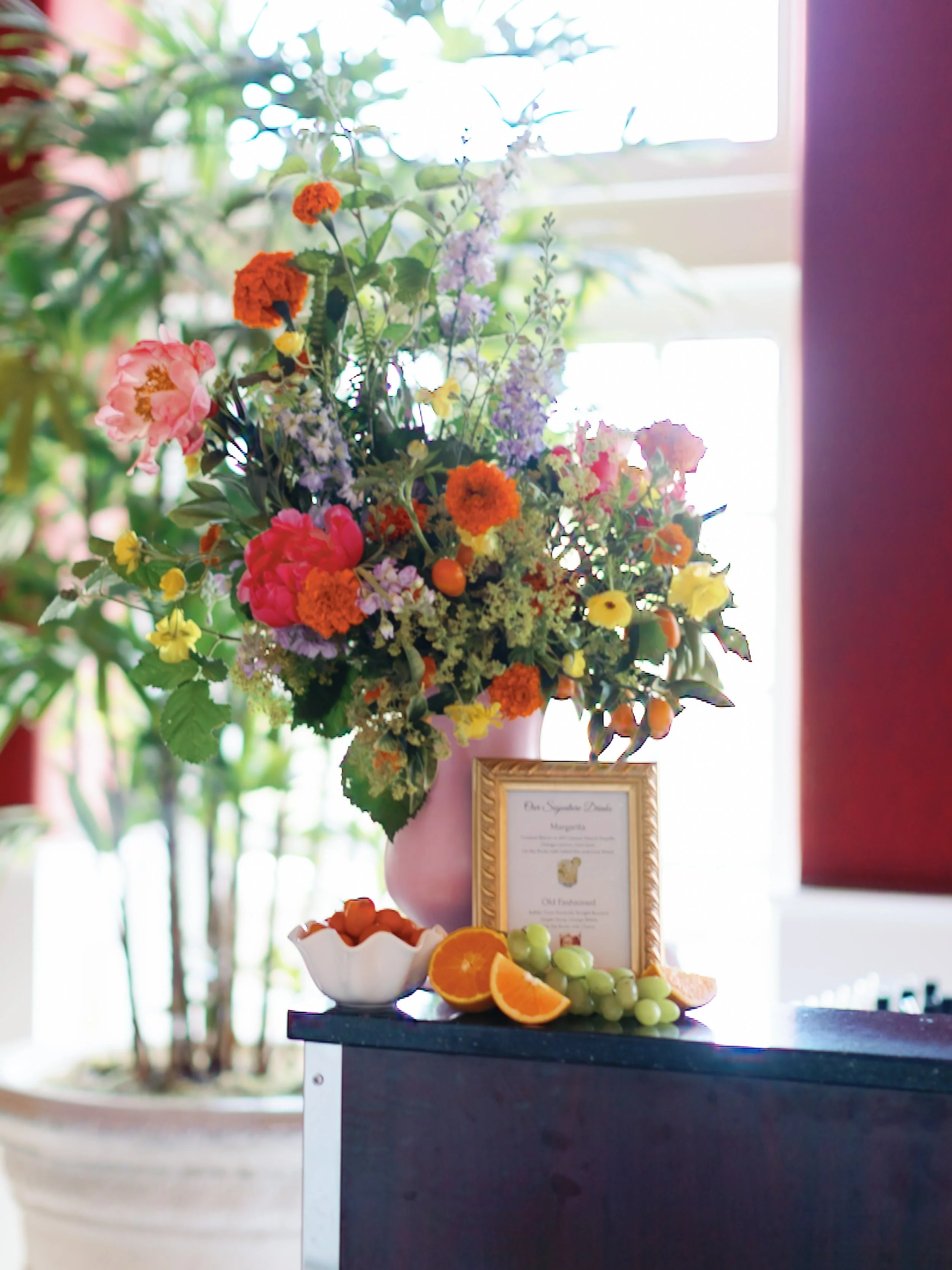 A large colorful flower arrangement in a pink vase on a table, with a small framed sign, sliced oranges, green grapes, and a bowl of wrapped candies in front of it.