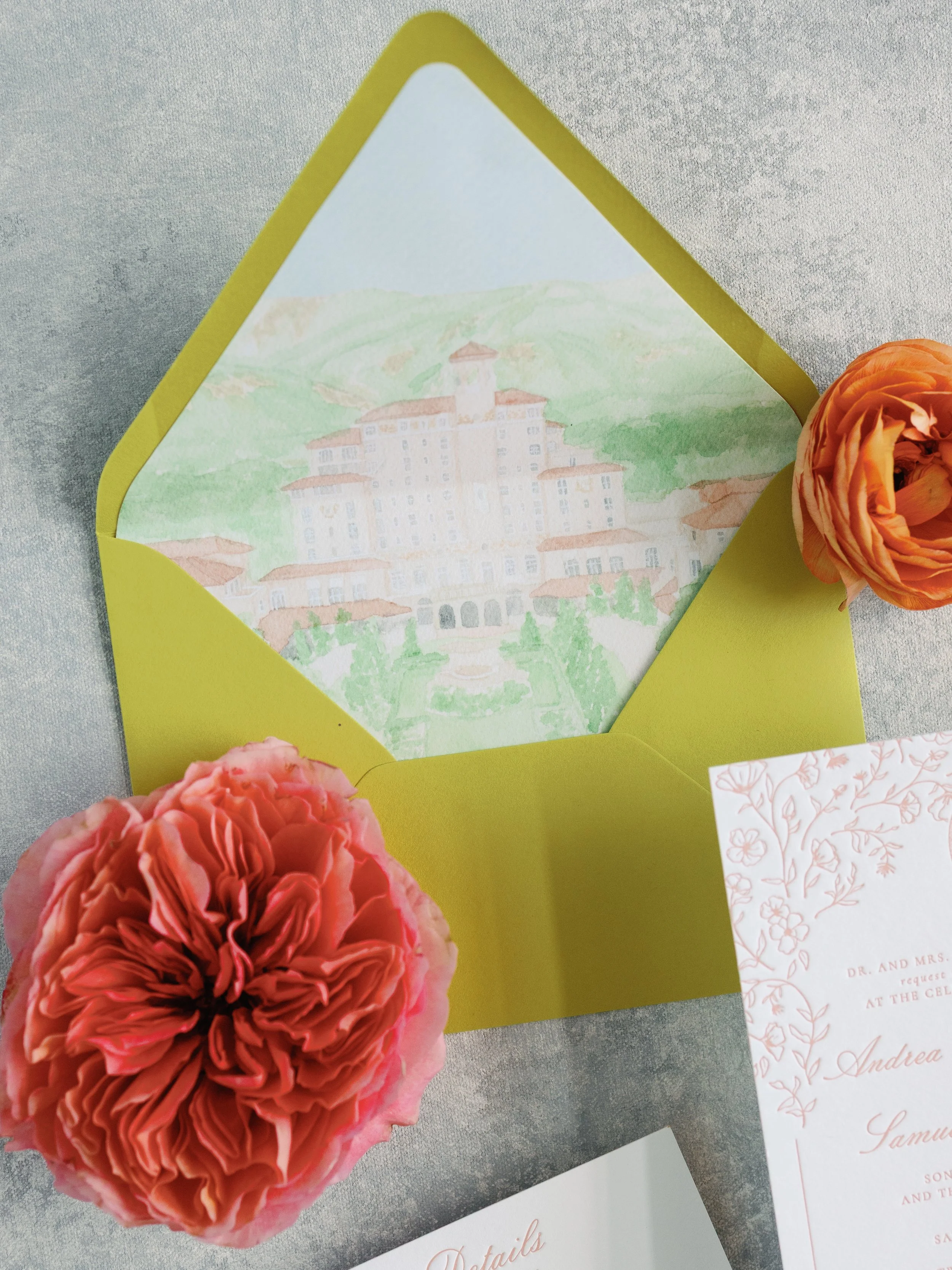 Open greeting card with watercolor painting of a castle on a green hillside, pink and orange flowers around it, on a gray surface with Pink floral invitation and white paper.