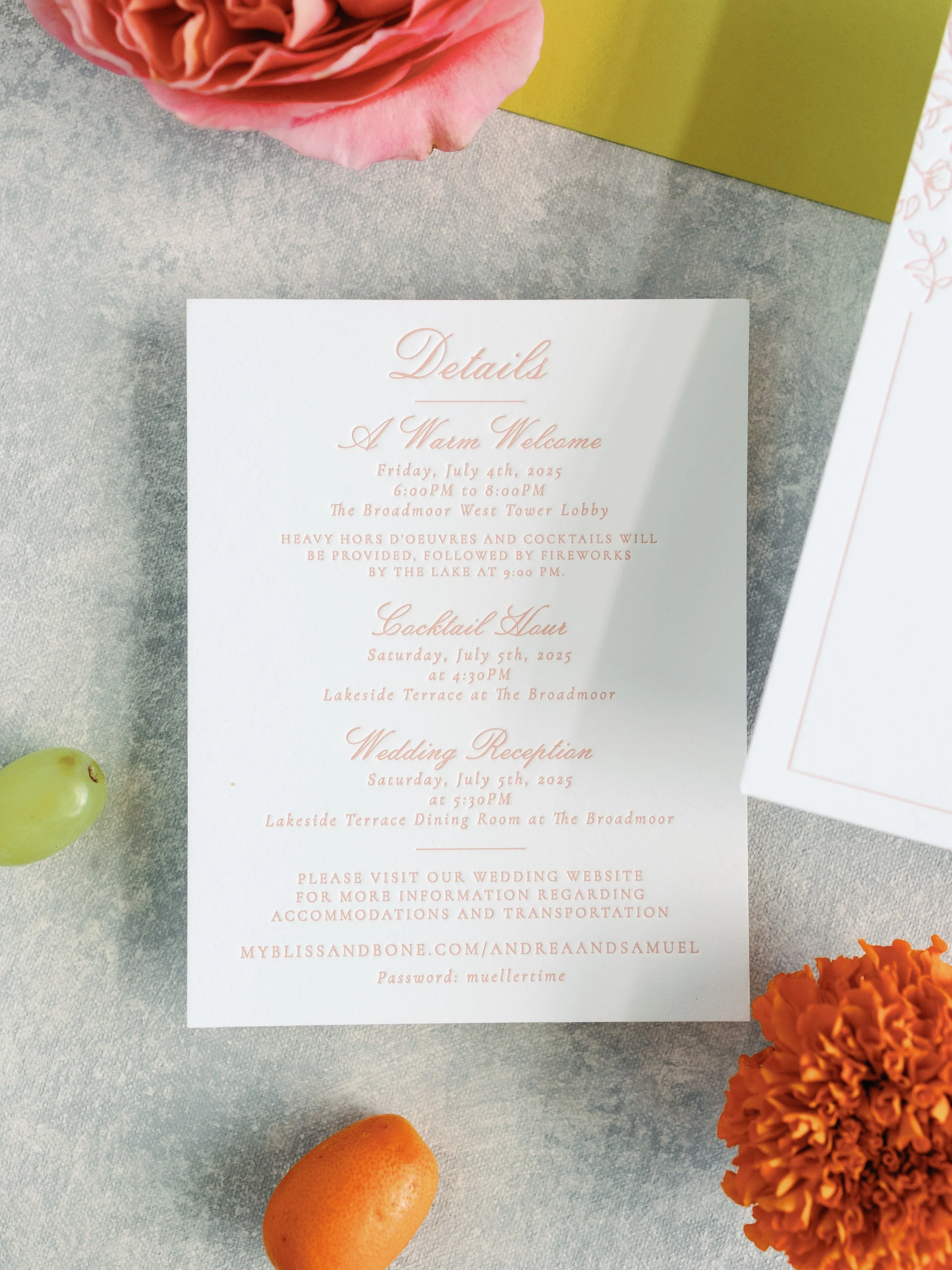 Wedding details card on table surrounded by orange flower, green grape, orange citrus, and pink flower.