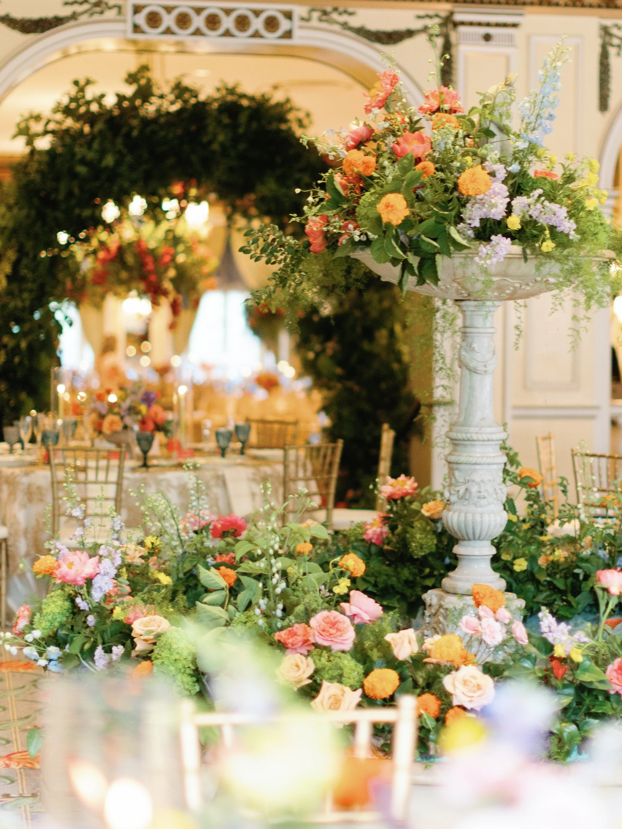 An elegant decorated event space with floral arrangements and tables set for a celebration, featuring large columns and a floral arch in the background.