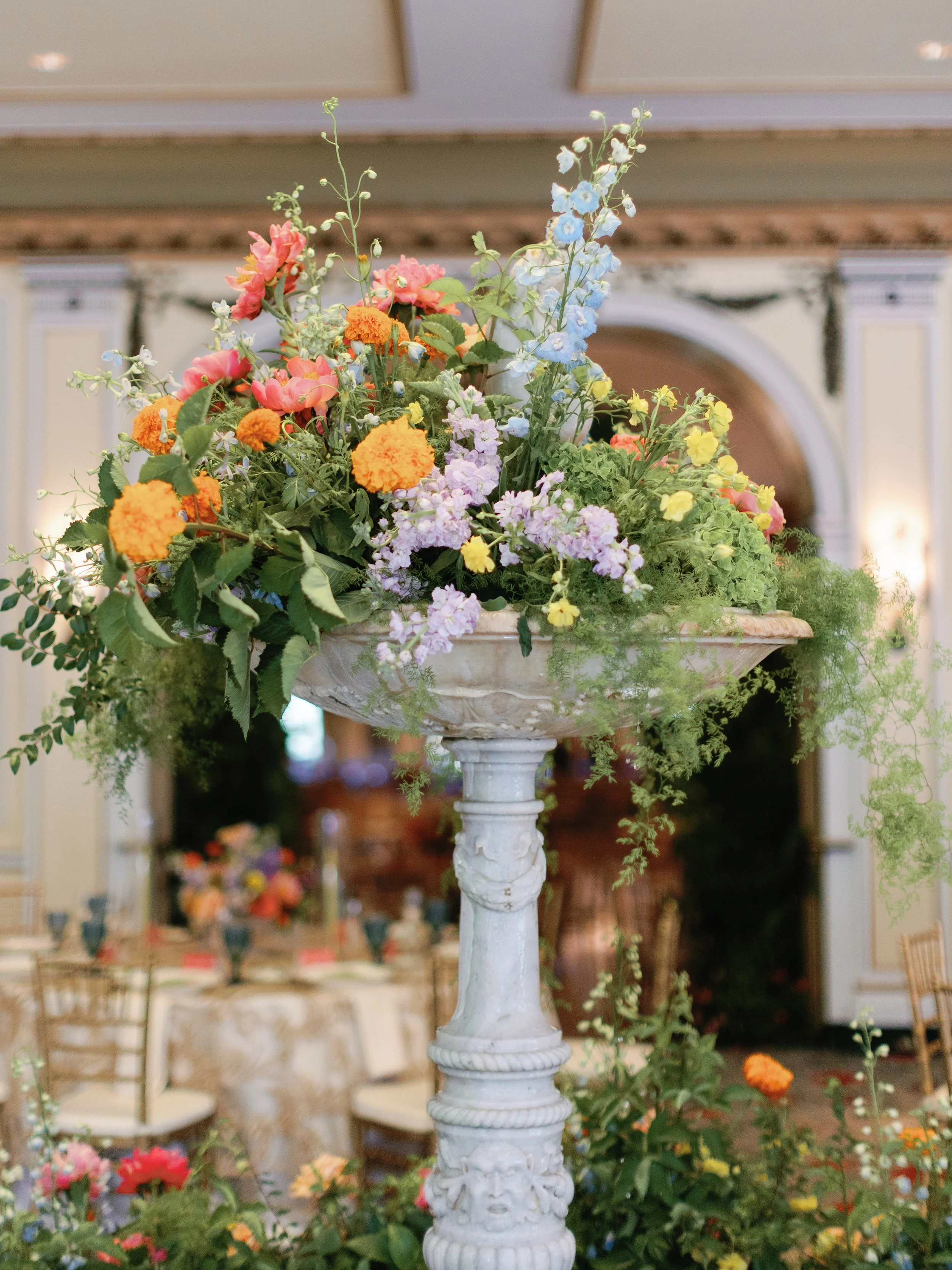 A large floral arrangement in a white pedestal vase filled with pink, orange, purple, blue, and yellow flowers, set in an elegant room with cream-colored walls and gold accents.