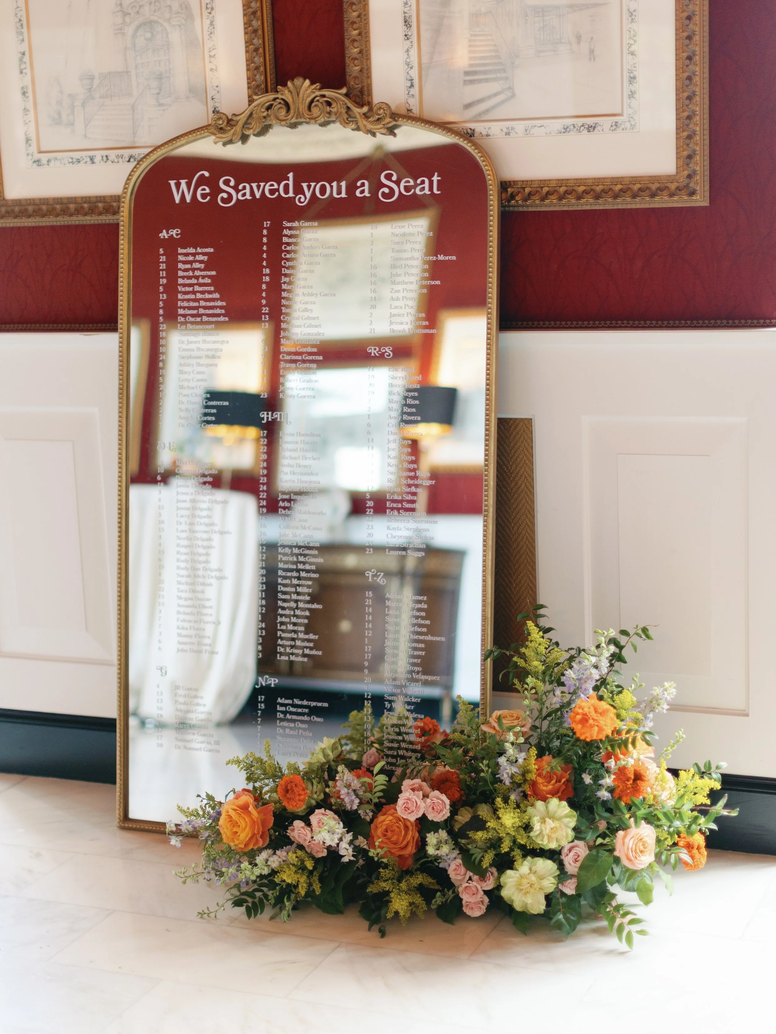 A decorative hotel or theater seating chart mirror with the title "We Saved You a Seat" at the top, surrounded by ornate gold detailing, with a colorful floral arrangement of roses, carnations, and other flowers at its base. The mirror reflects the i