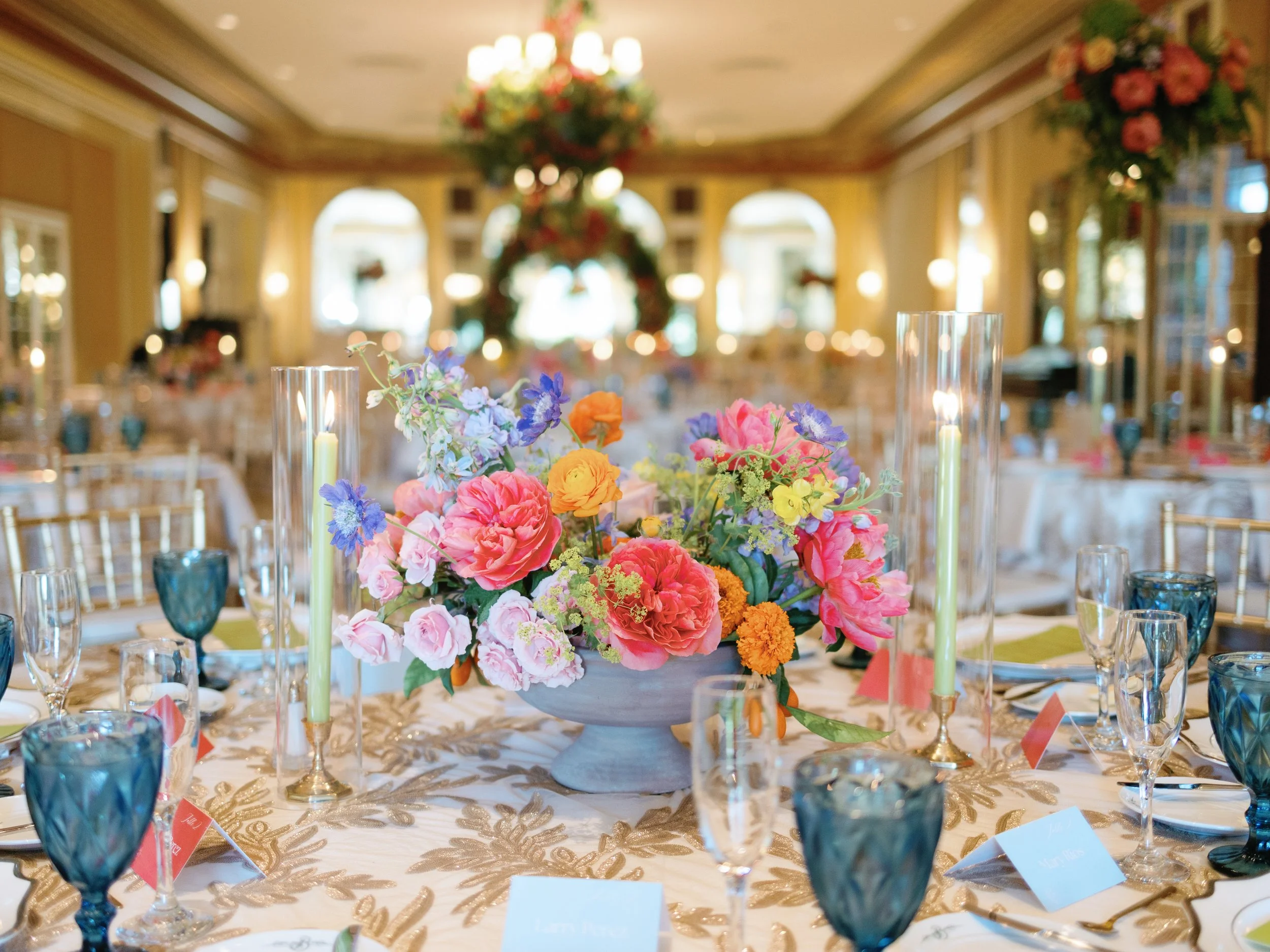 A banquet table decorated with a pink, purple, and yellow floral centerpiece, surrounded by wine glasses, candles, and place cards, set in an elegant room with large windows and floral arrangements.