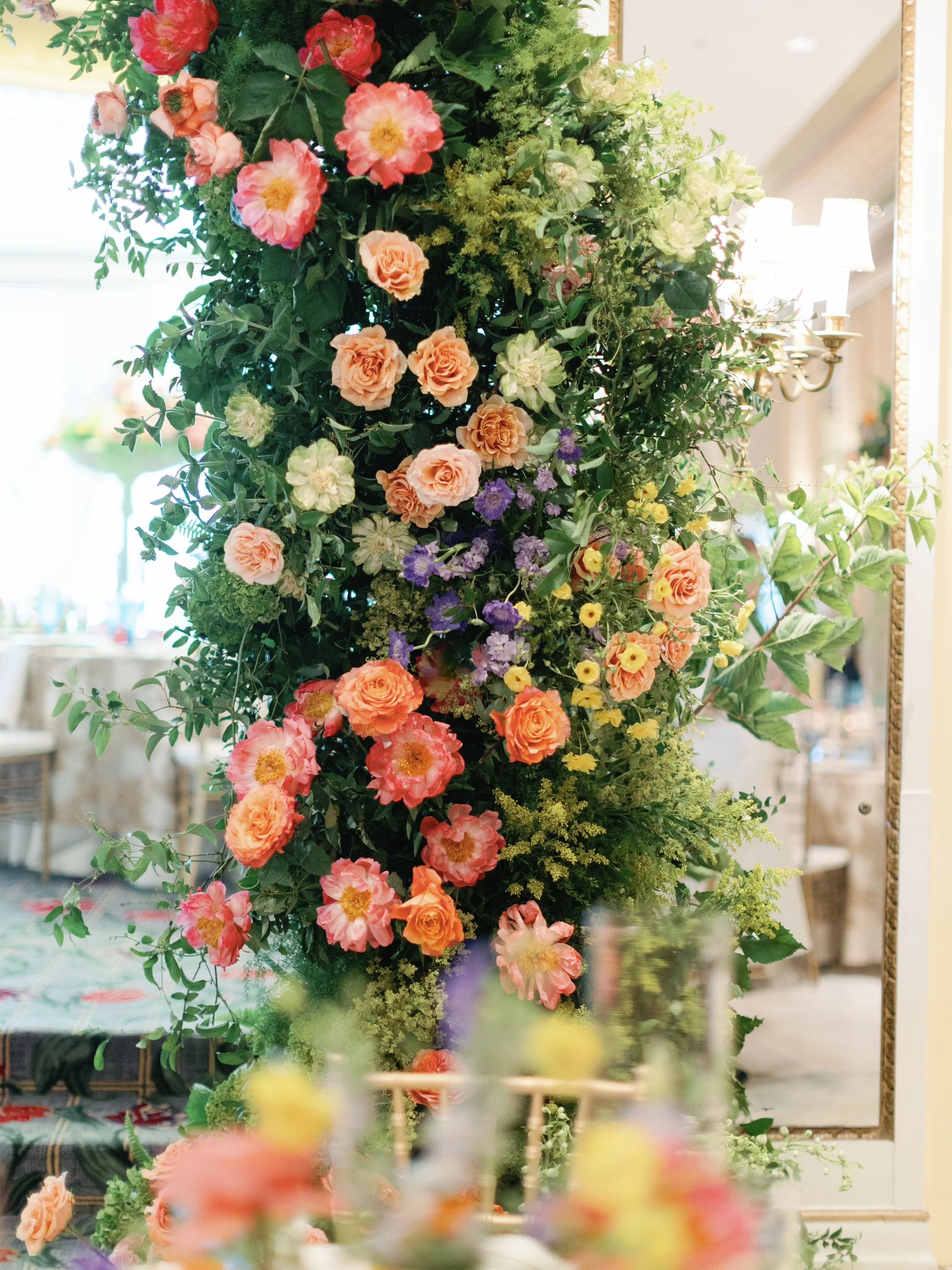 Large floral arrangement with pink, peach, purple, and white flowers, surrounded by green leaves, displayed indoors near a mirror and a chandelier.