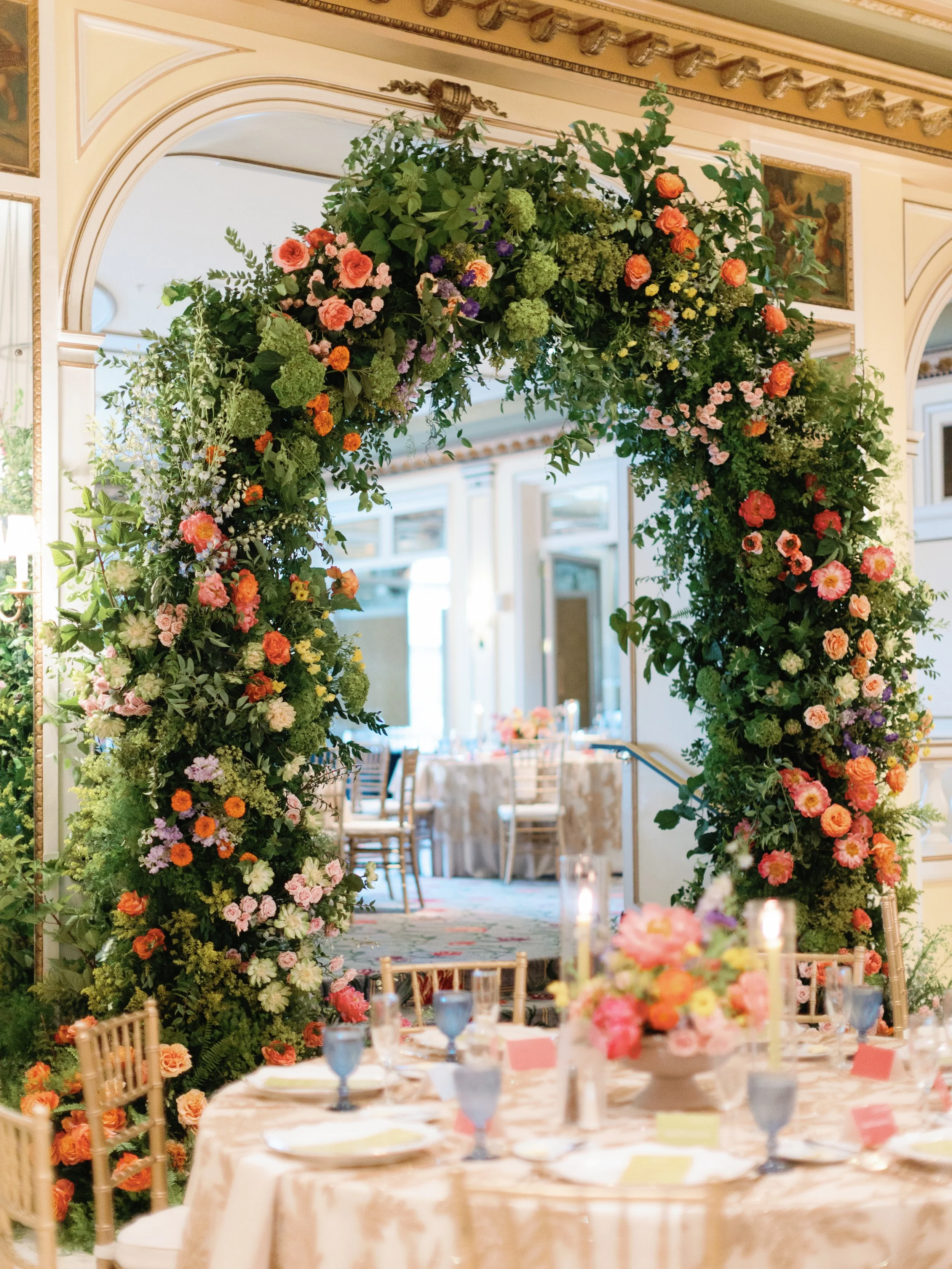 A floral arch with pink, peach, purple, and green flowers decorates a banquet hall entrance for a wedding or event. In the foreground, a round table is set with pink and blue glassware, plates, and a floral centerpiece.