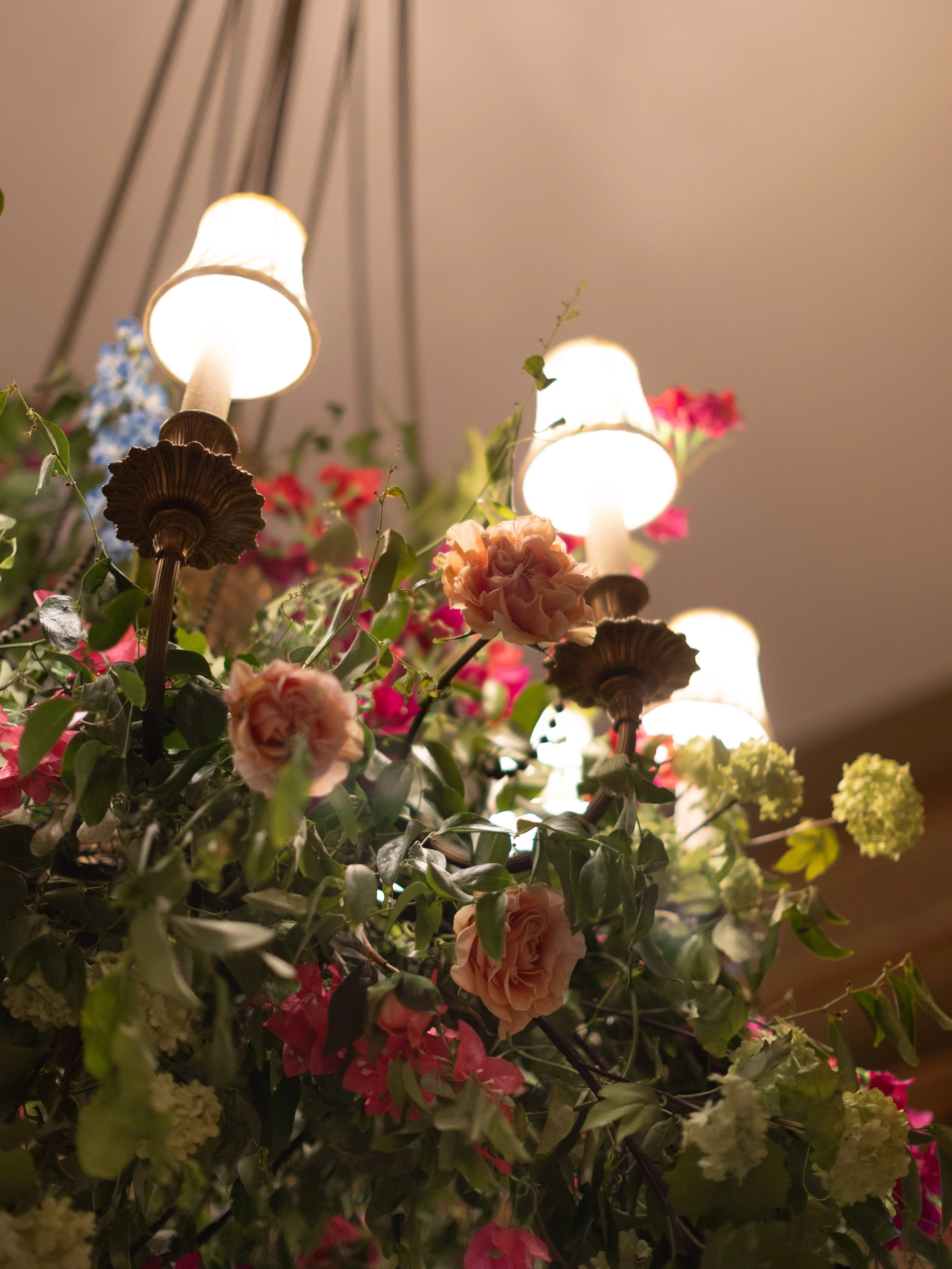 A chandelier with five lit lamps surrounded by pink, white, and blue flowers, with green foliage underneath.