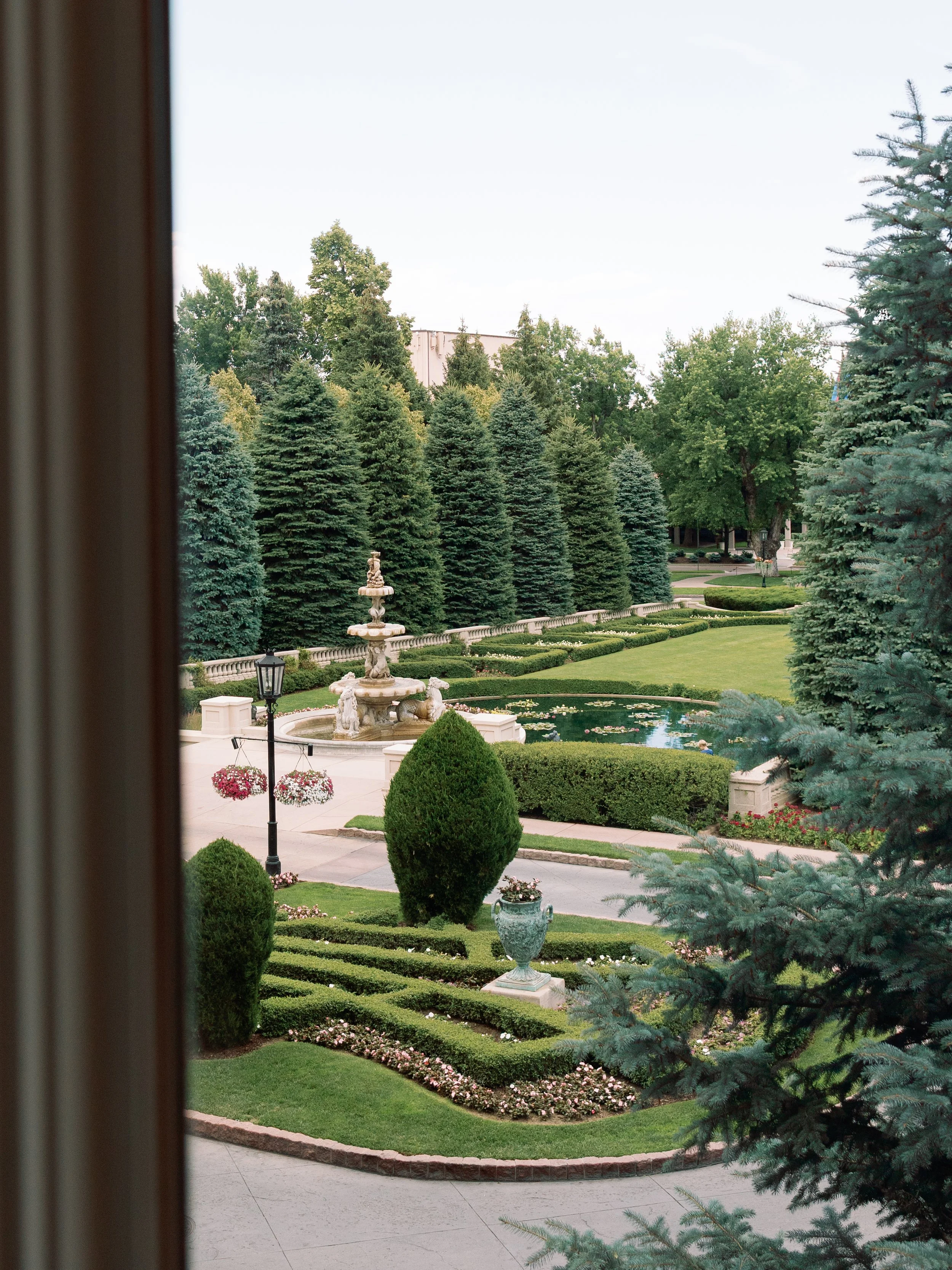 View of a well-maintained garden with a fountain, trimmed bushes, flowerbeds, a pond, and tall evergreen trees, seen through a window.
