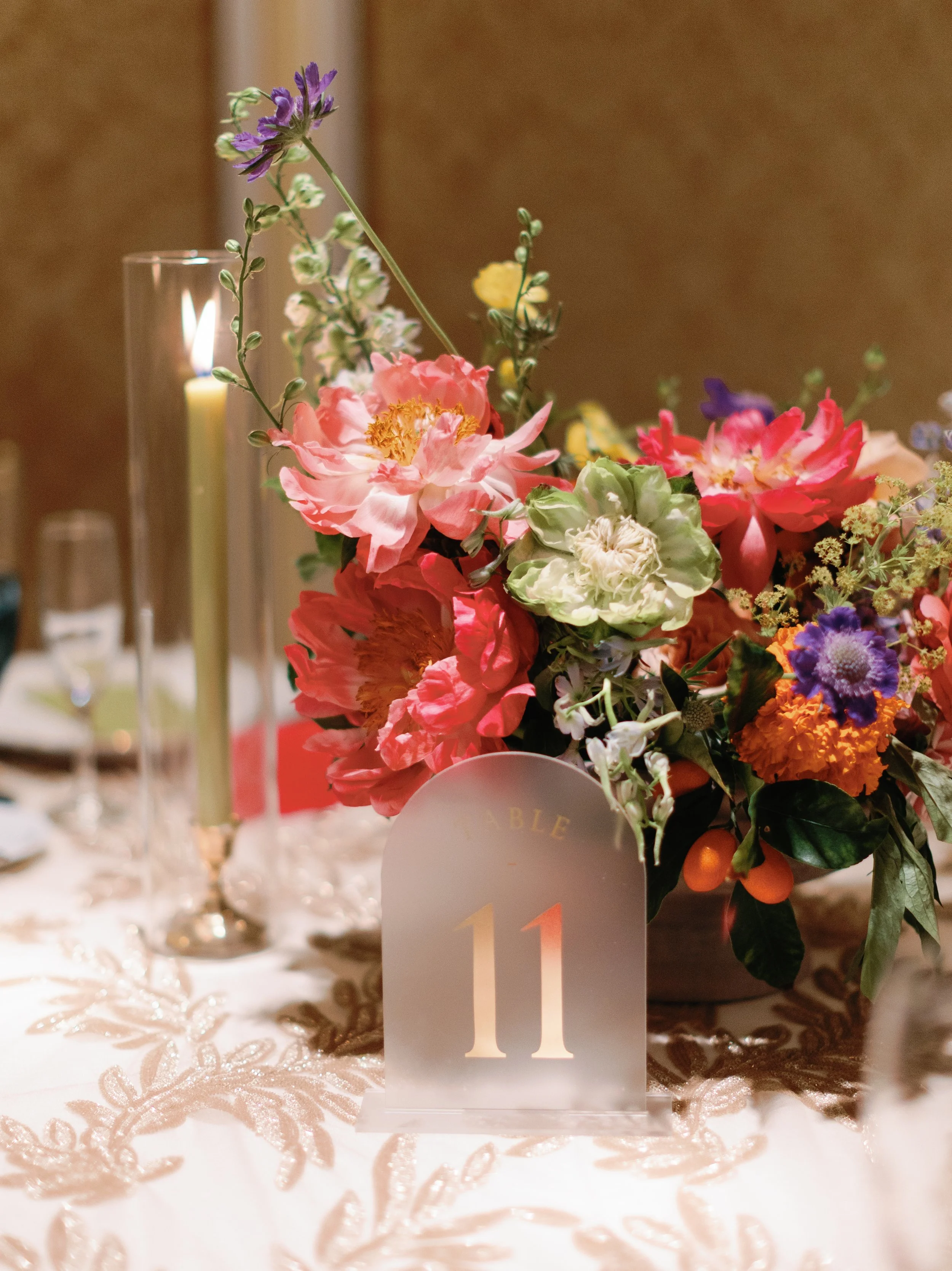 A floral centerpiece with pink, purple, white, orange, and yellow flowers on a table with a table number 11 in front, a lit candle in a glass holder, and elegant table setting.