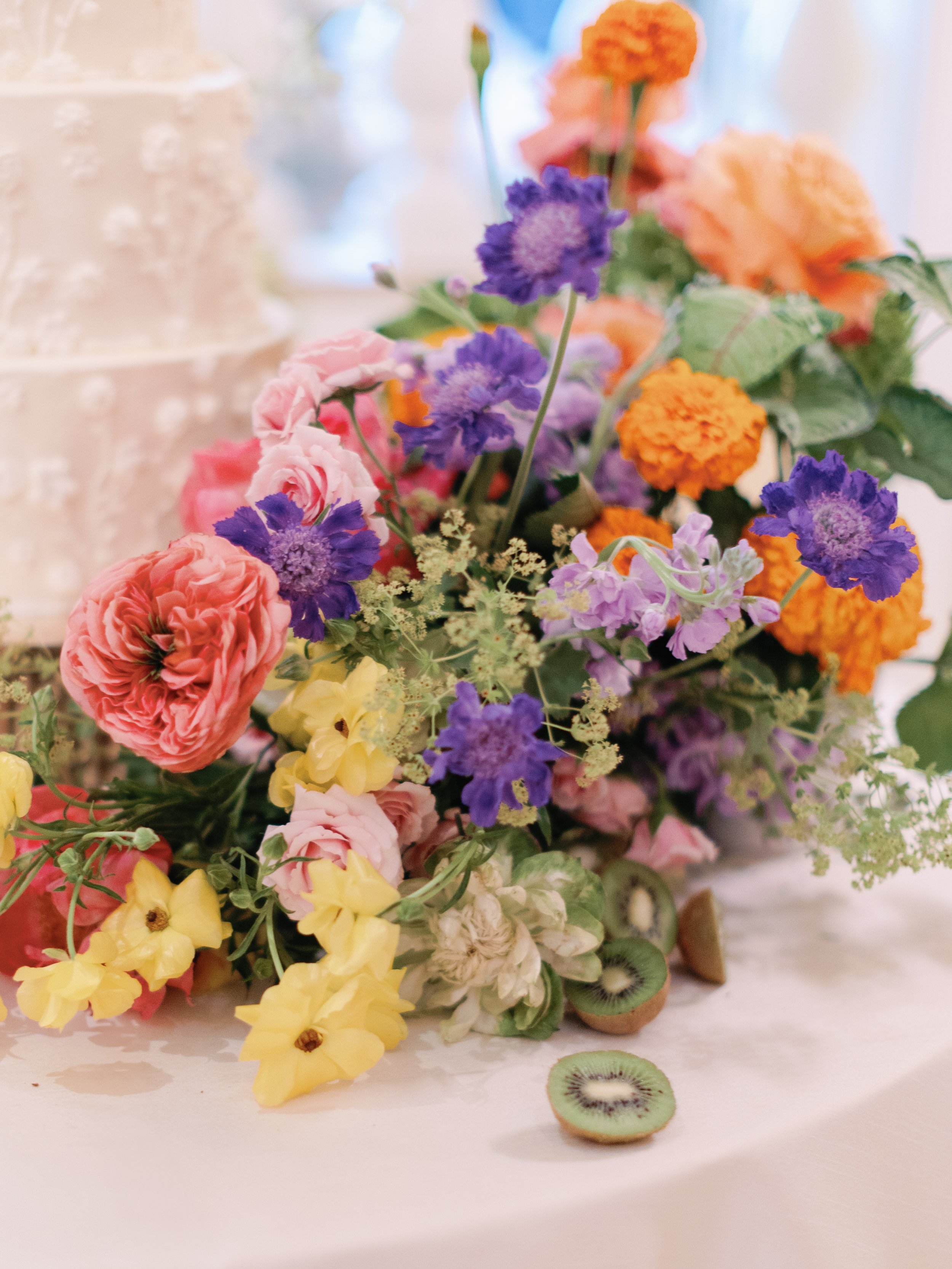 Colorful bouquet of pink, purple, yellow, and orange flowers with a sliced kiwi fruit in front.