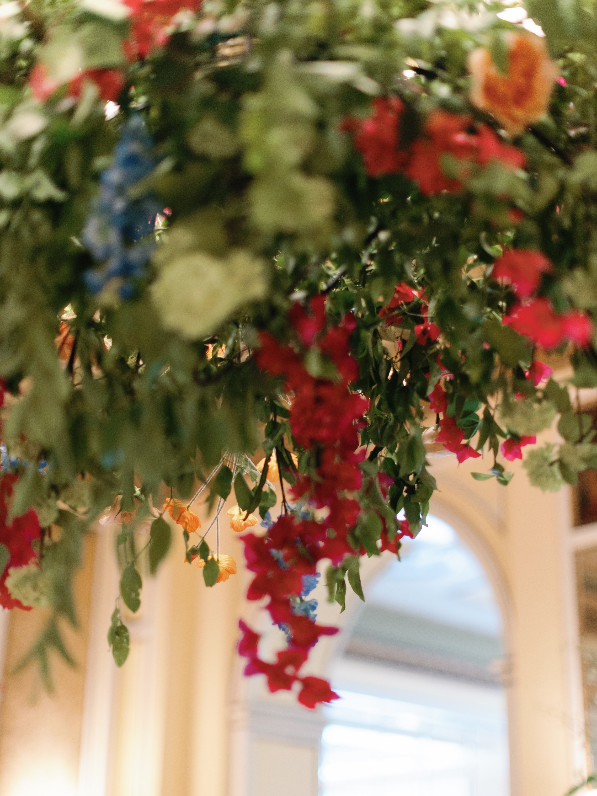 Hanging arrangement of colorful flowers, including red, orange, yellow, blue, and white blooms, inside a building with a window in the background.