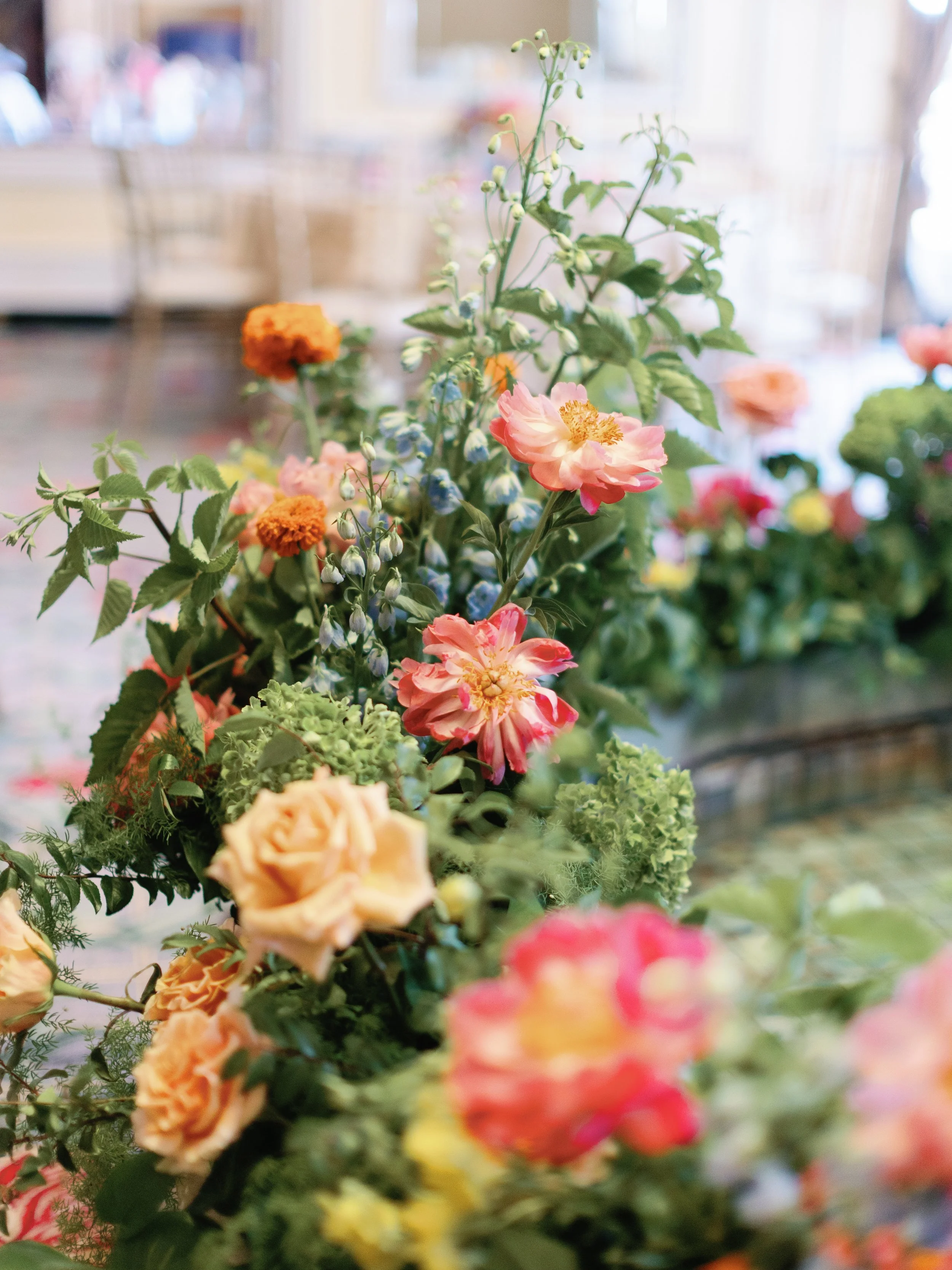 A colorful bouquet of various flowers, including pink, peach, orange, and blue blossoms, arranged with green foliage in an indoor setting.