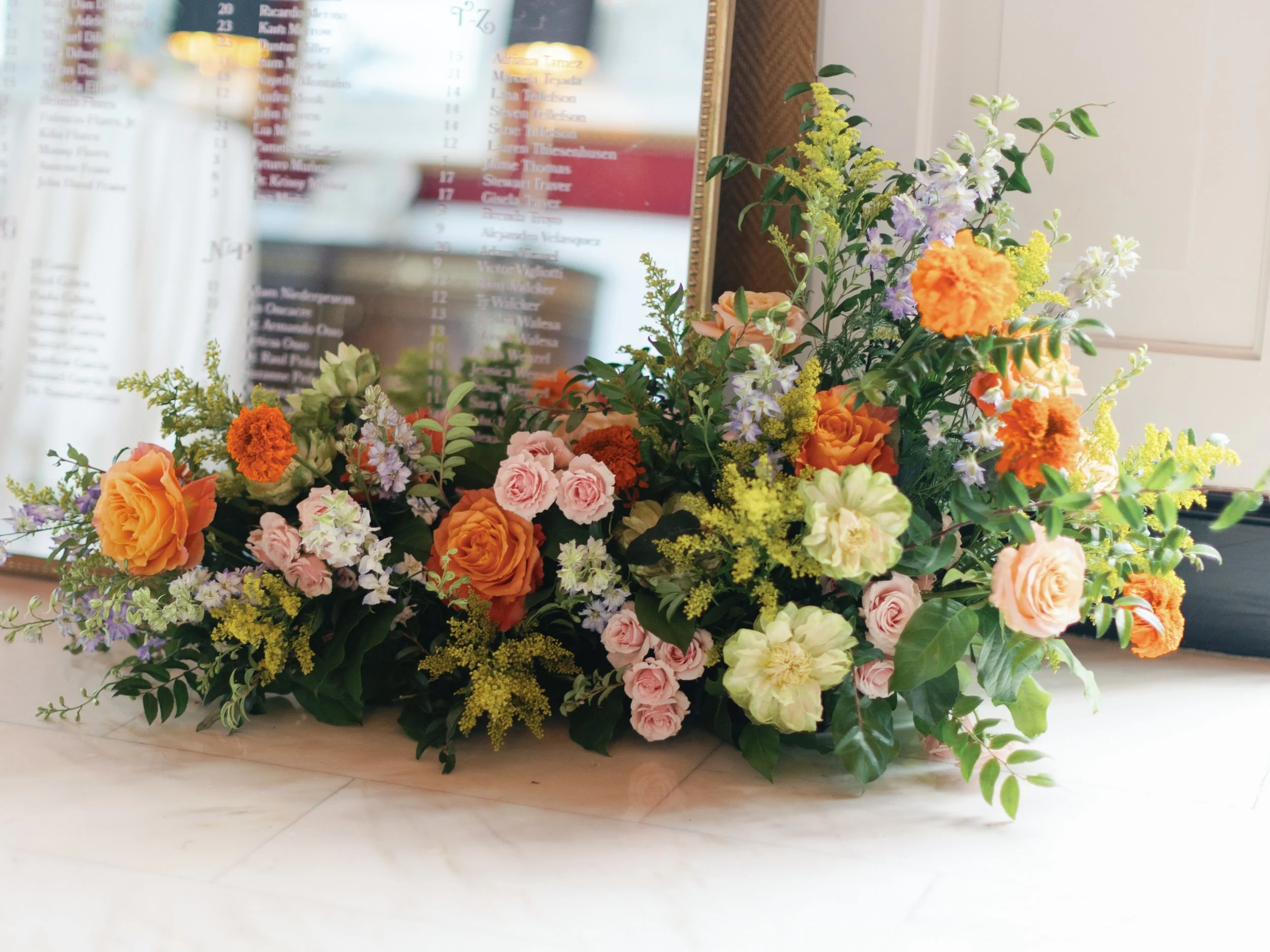 A colorful arrangement of flowers with orange, pink, yellow, and purple blooms, with green foliage, positioned on a marble surface indoor, in front of a mirror or glass with a blurred background.
