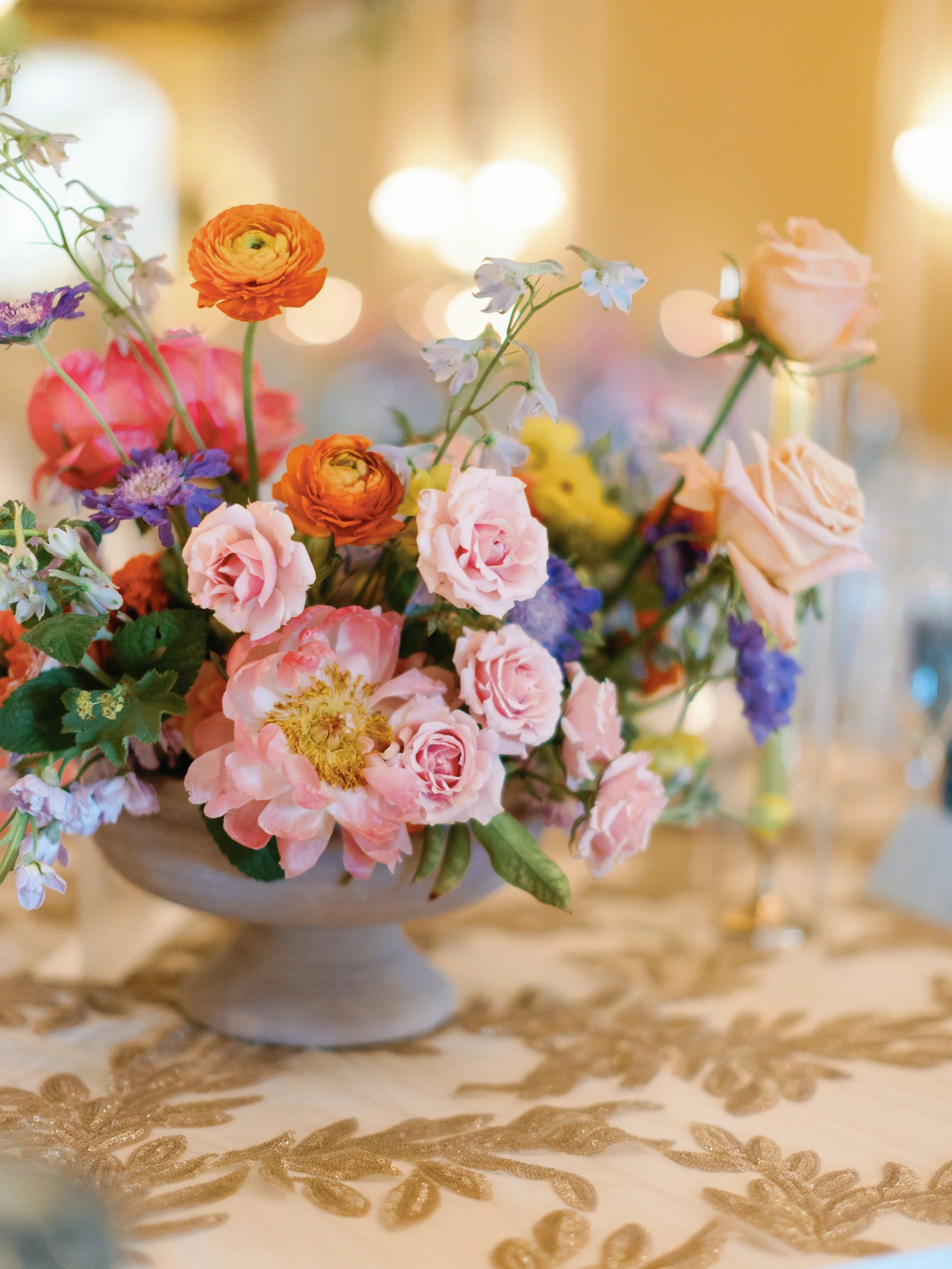 A colorful bouquet of flowers including pink roses, orange ranunculus, purple blooms, and small white and blue flowers in a white vase on a decorative tablecloth.