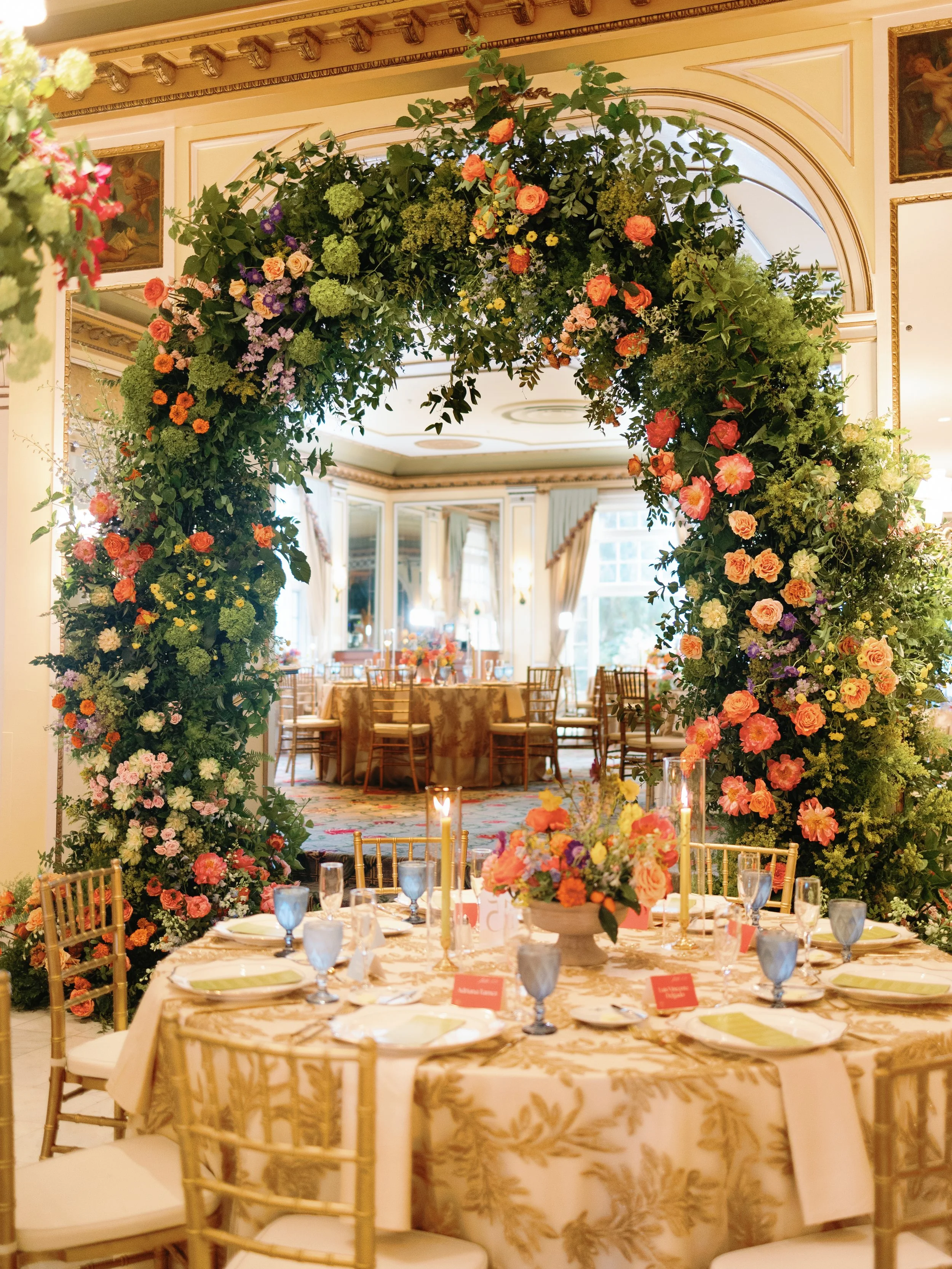 Elegant dining room with round table, floral centerpiece, gold chairs, and a floral arch in a luxurious banquet hall.