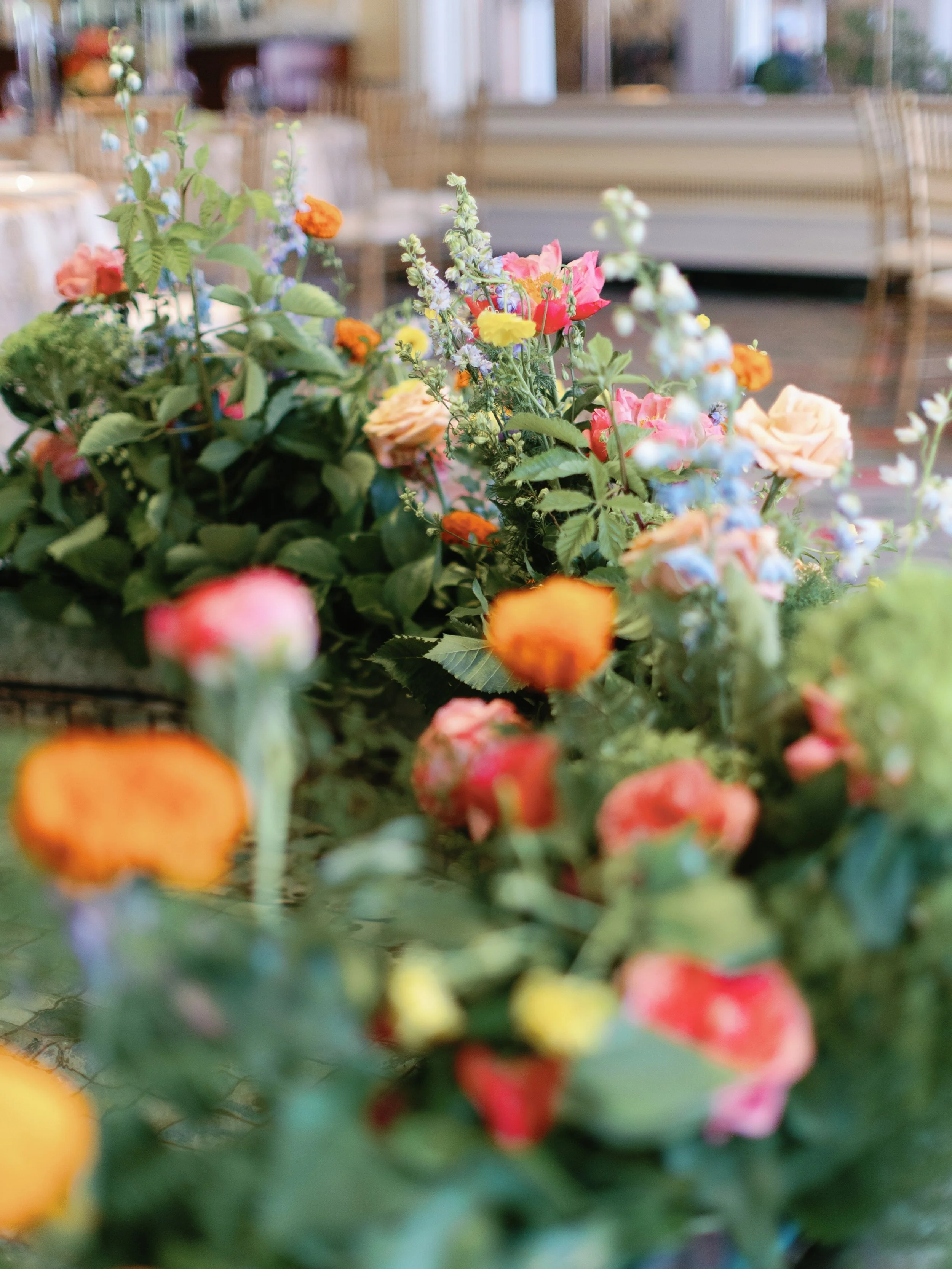 Close-up of a colorful flower arrangement with pink, orange, and blue flowers and green leaves, set in a blurred indoor setting.