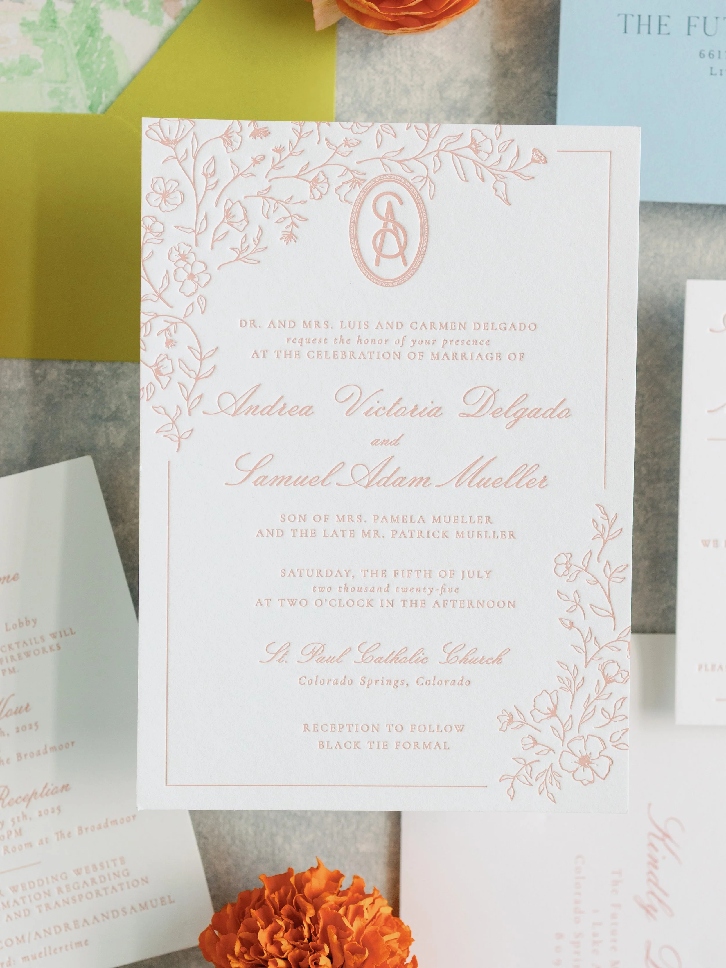 A wedding invitation card with floral embellishments and elegant script, laid on a table with orange flowers and other stationery.
