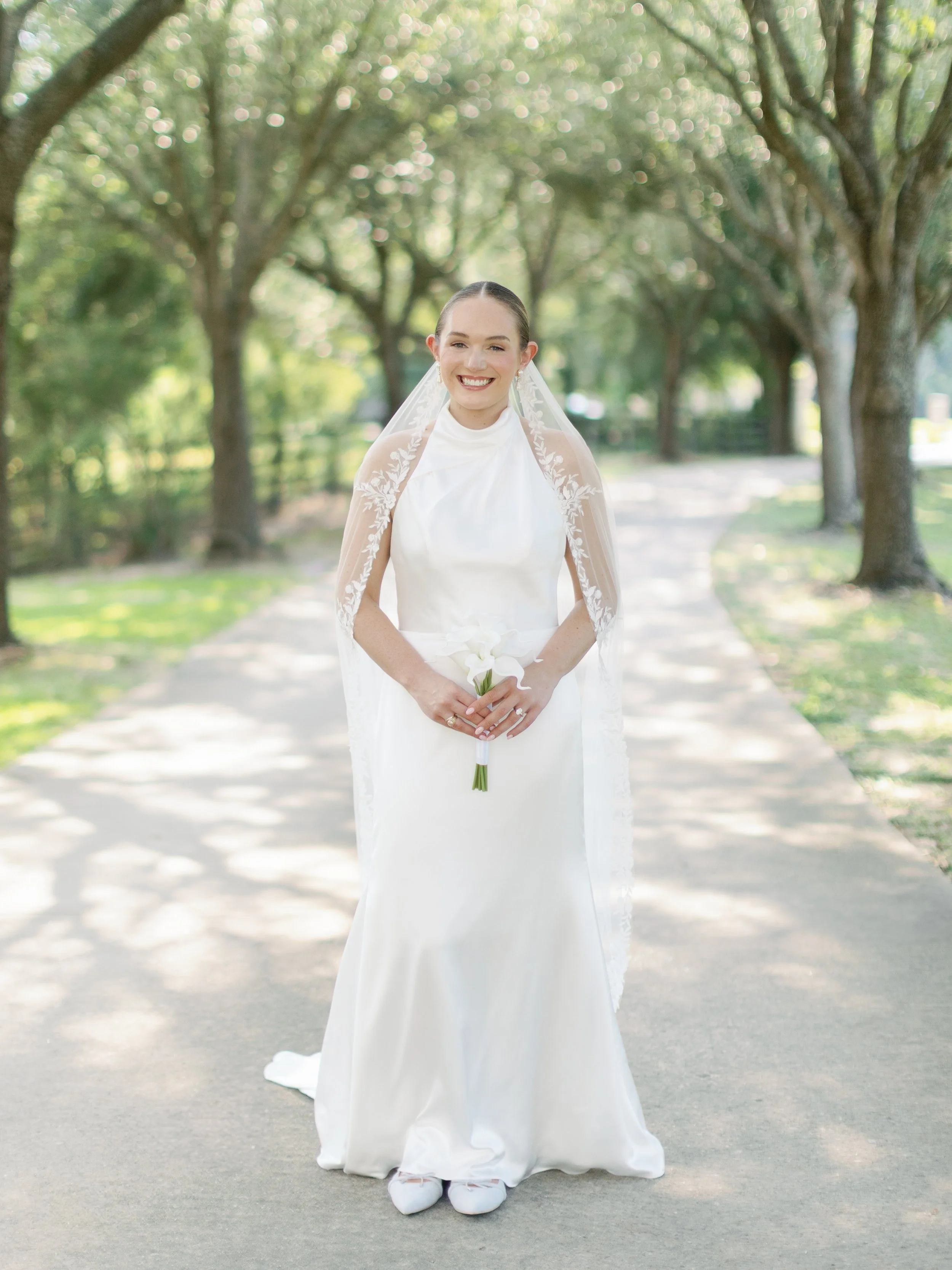 A bride in a white wedding dress and veil stands on a tree-lined path, smiling and holding a small bouquet of white flowers.