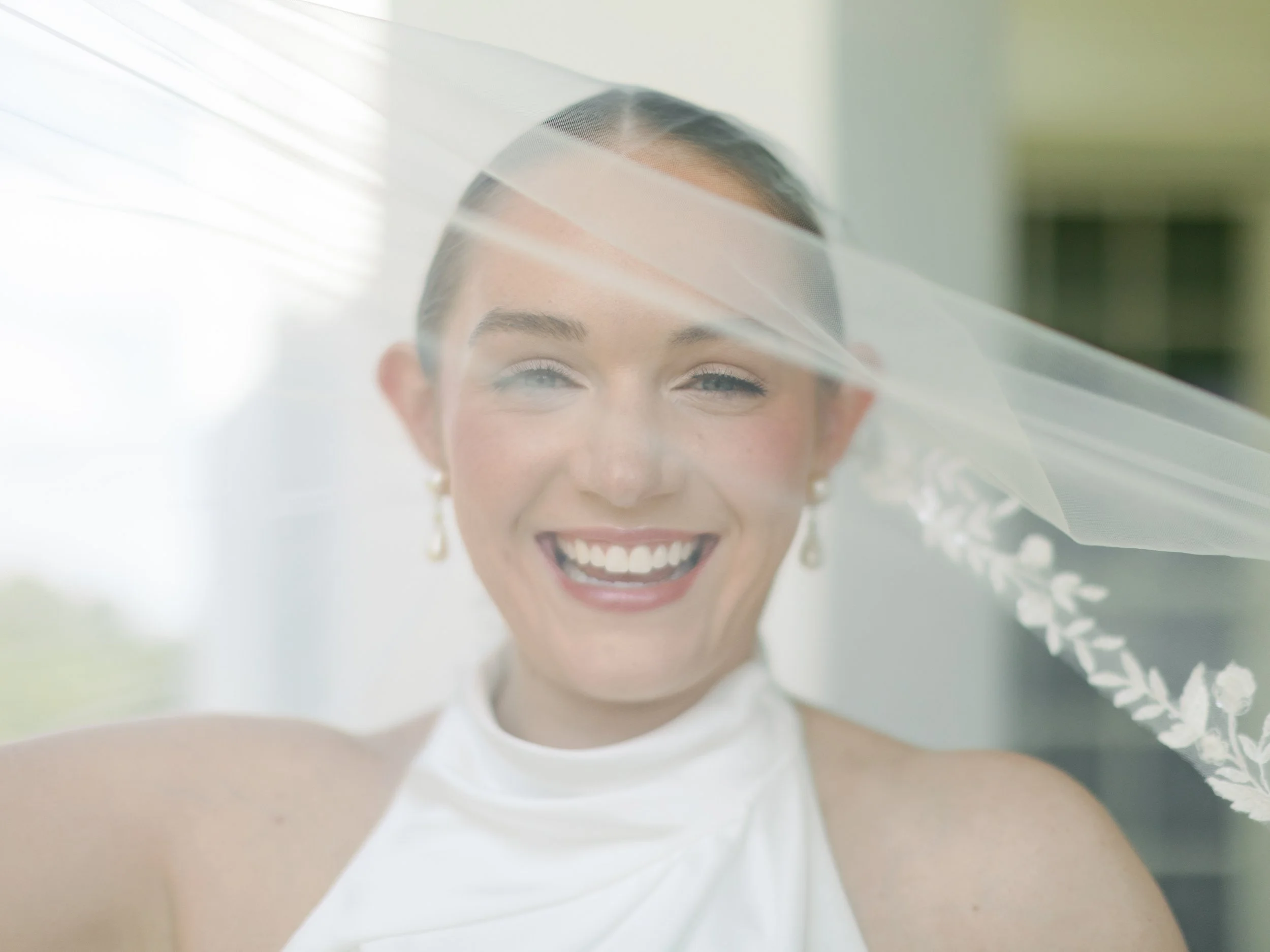 A smiling woman with a wedding veil and earrings, wearing a white dress, indoors with sunlight streaming through a window.