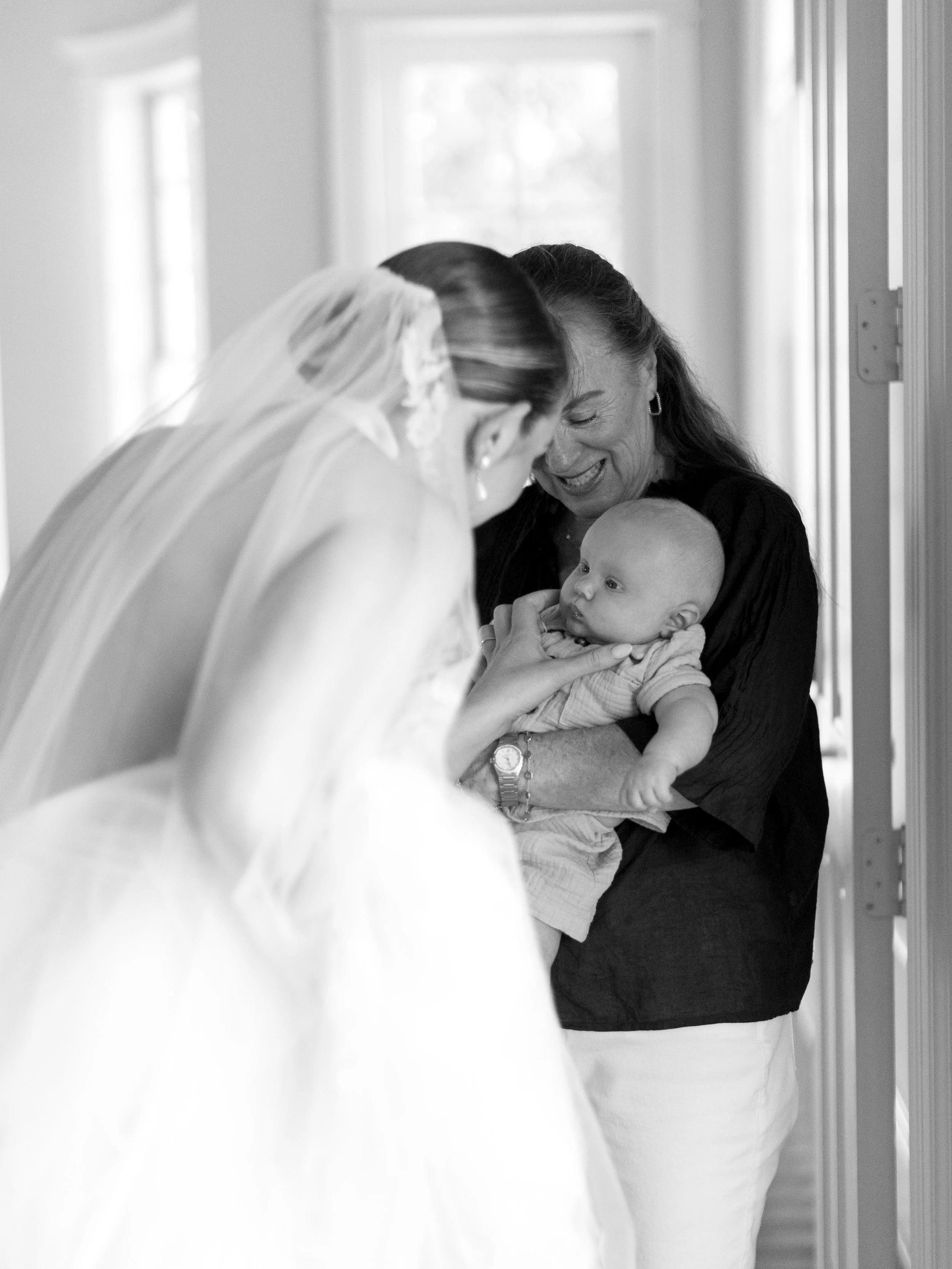 A woman holding a baby while a bride in a wedding gown and veil leans in smiling.