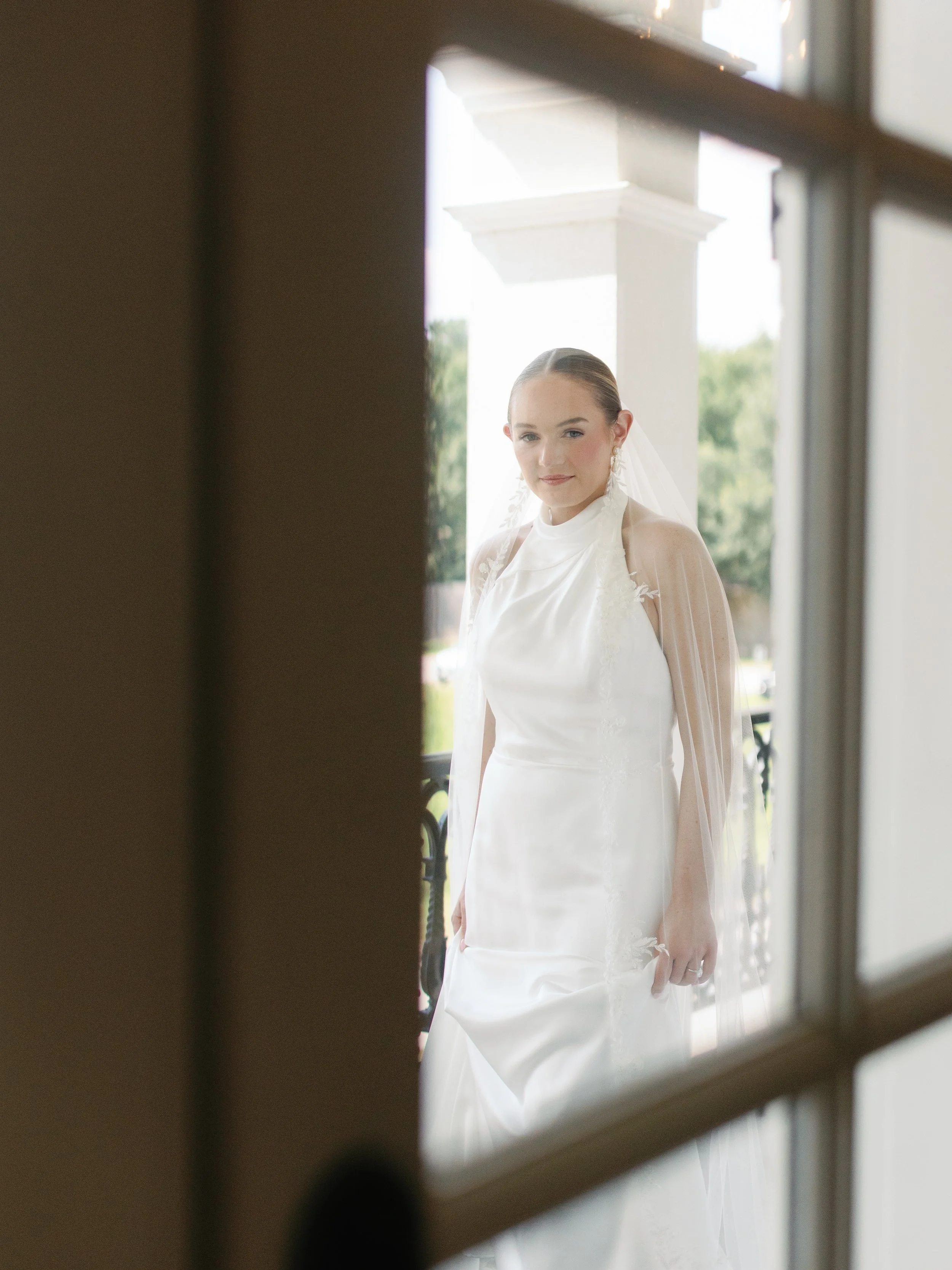 A bride in a white wedding dress and veil is looking at the camera outside a house, framed by a window.