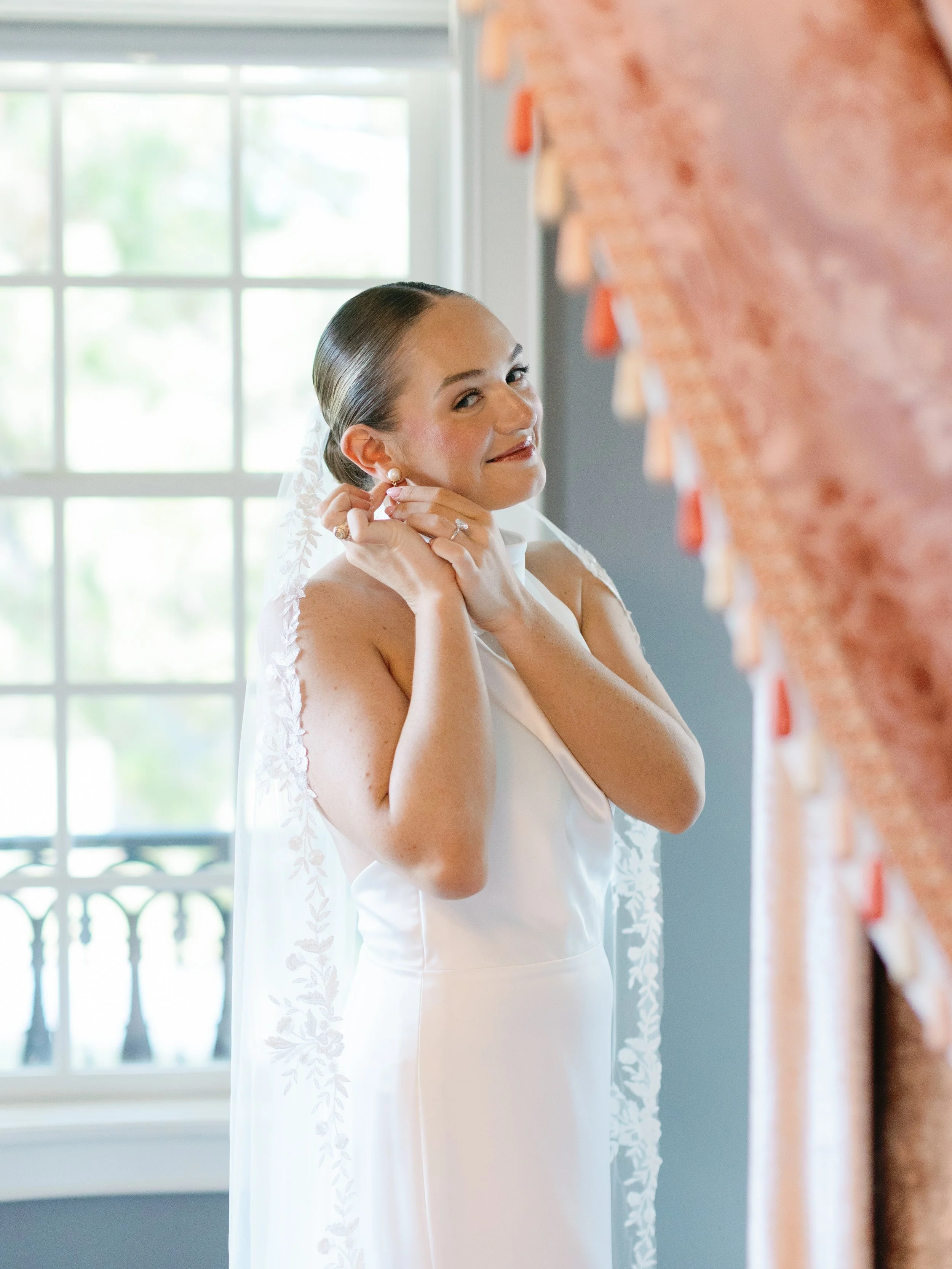 A bride in a white wedding dress and veil is adjusting her pearl earring in front of a window.