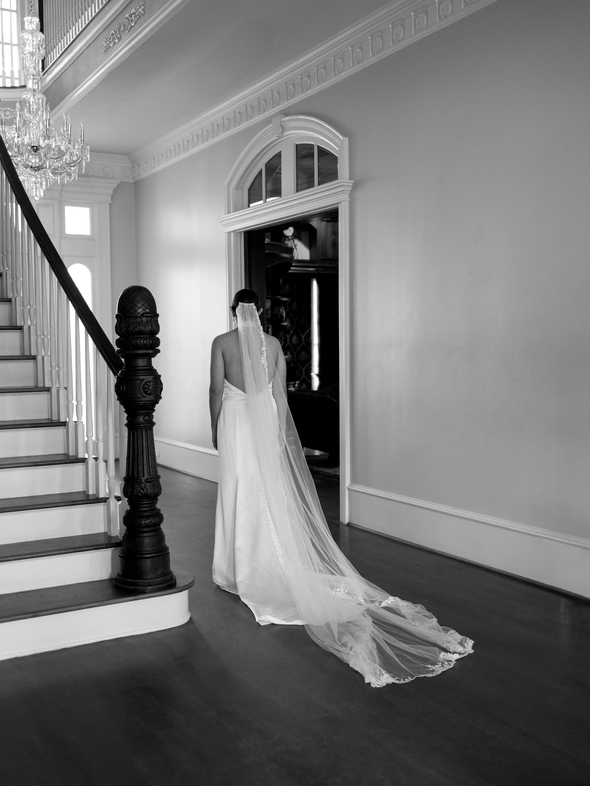 A bride in a wedding dress with a long veil stands in a grand, elegantly decorated hallway near a staircase and doorway.