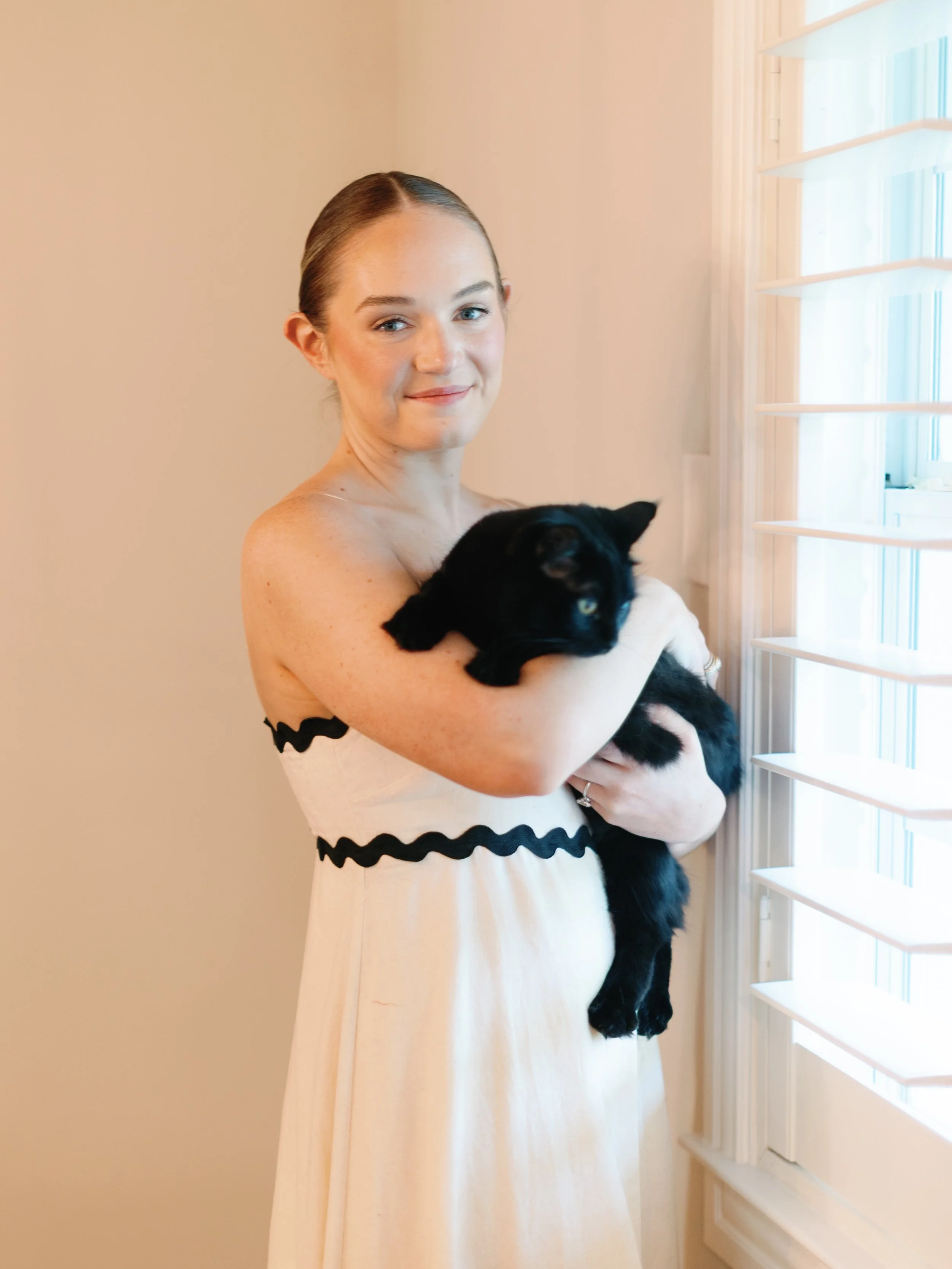 A woman with a bun hairstyle holding a black cat near a window with shutters, wearing a light-colored dress with black wavy trim.
