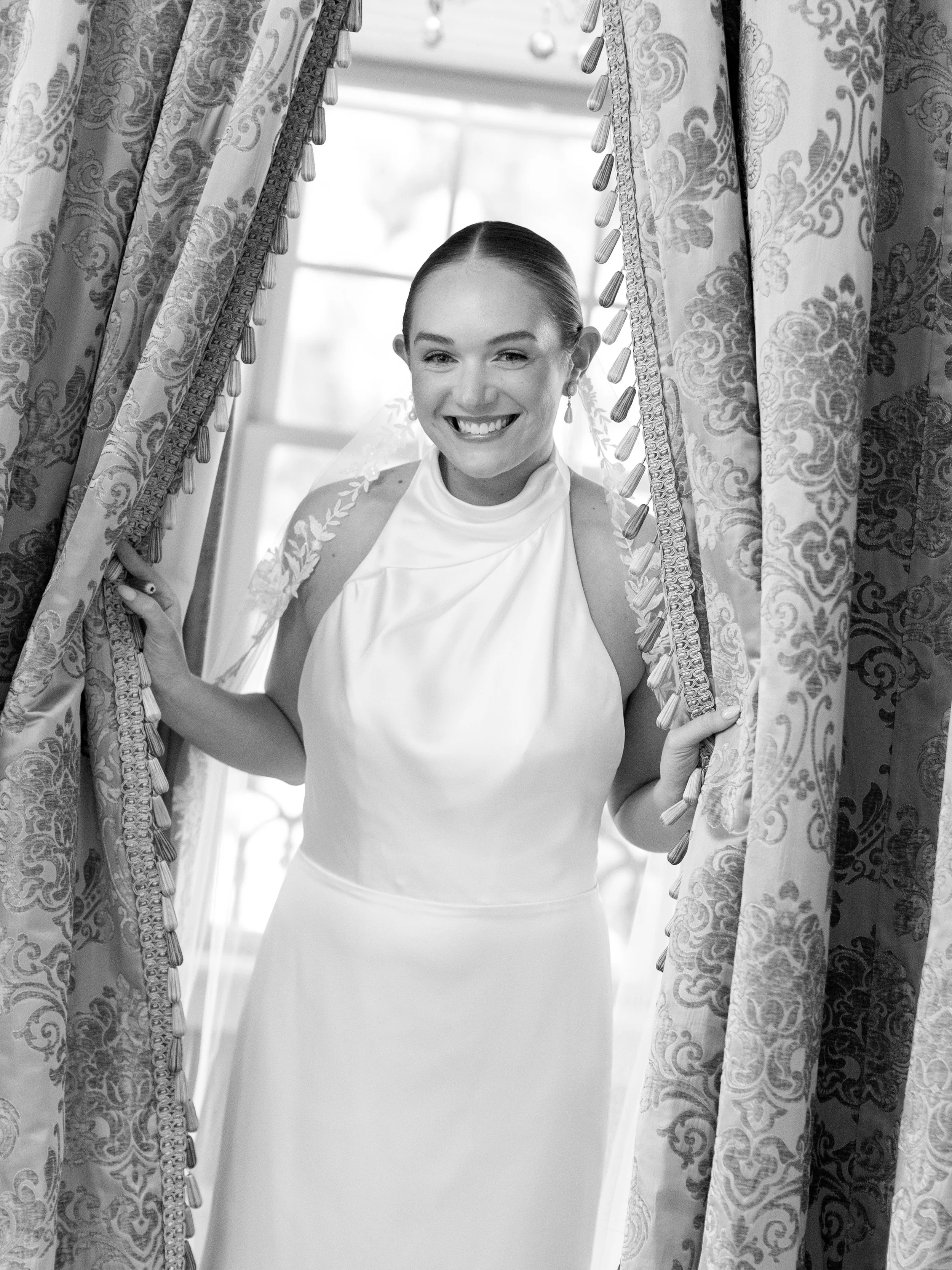 A smiling woman in a sleeveless white dress stands between curtain panels, holding them open, with a window in the background.