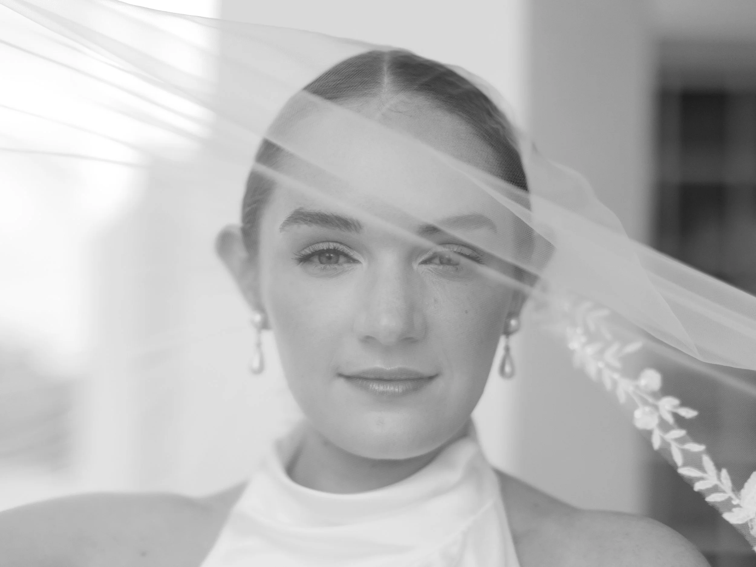 Black and white portrait of a woman with a veil covering part of her face, wearing earrings and a high-neck dress, indoors.